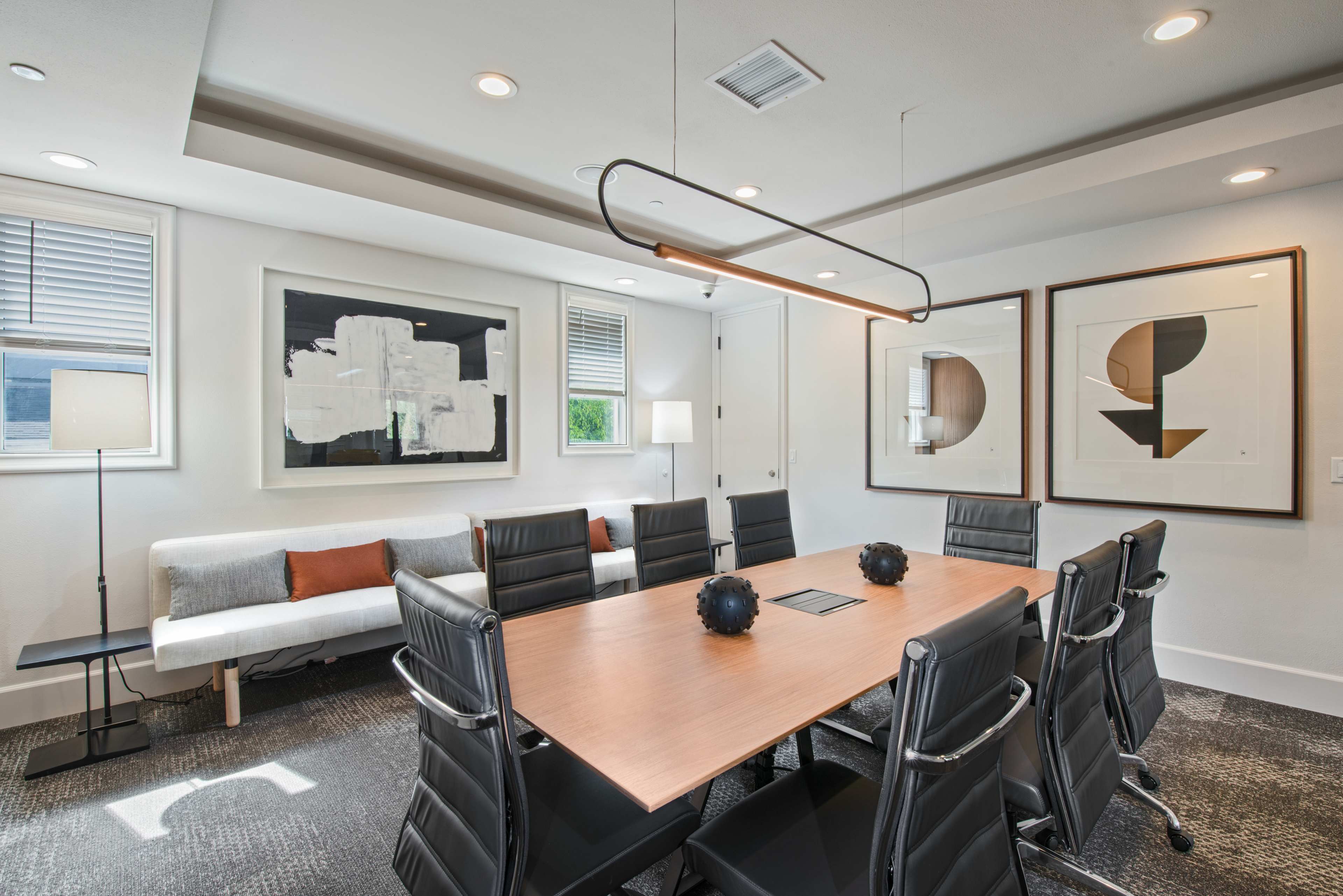 A modern conference room features a rectangular wooden table surrounded by six black leather chairs, with abstract artwork on the walls and a sofa along one side.