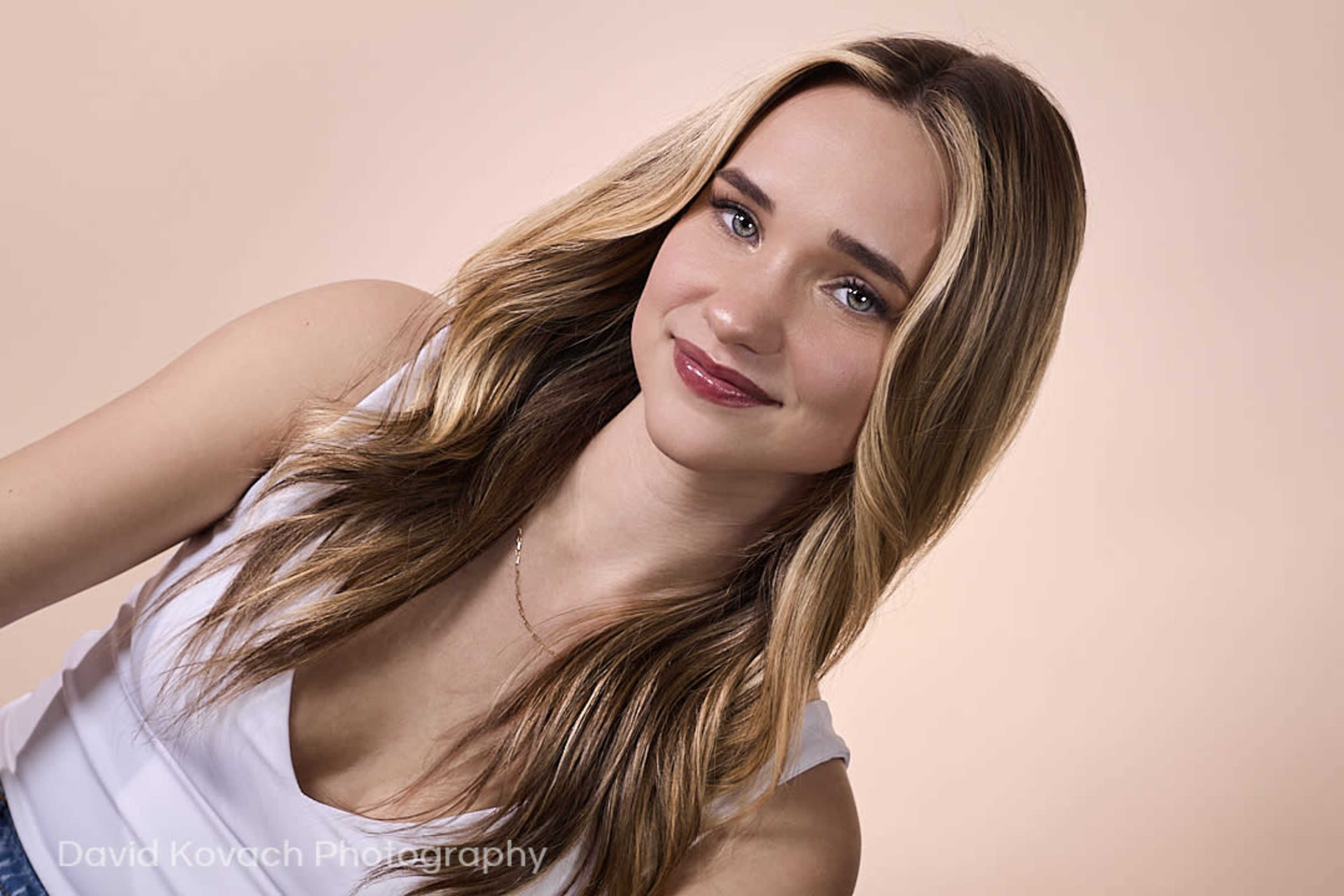 A young woman with long, wavy hair is smiling against a light, neutral background.
