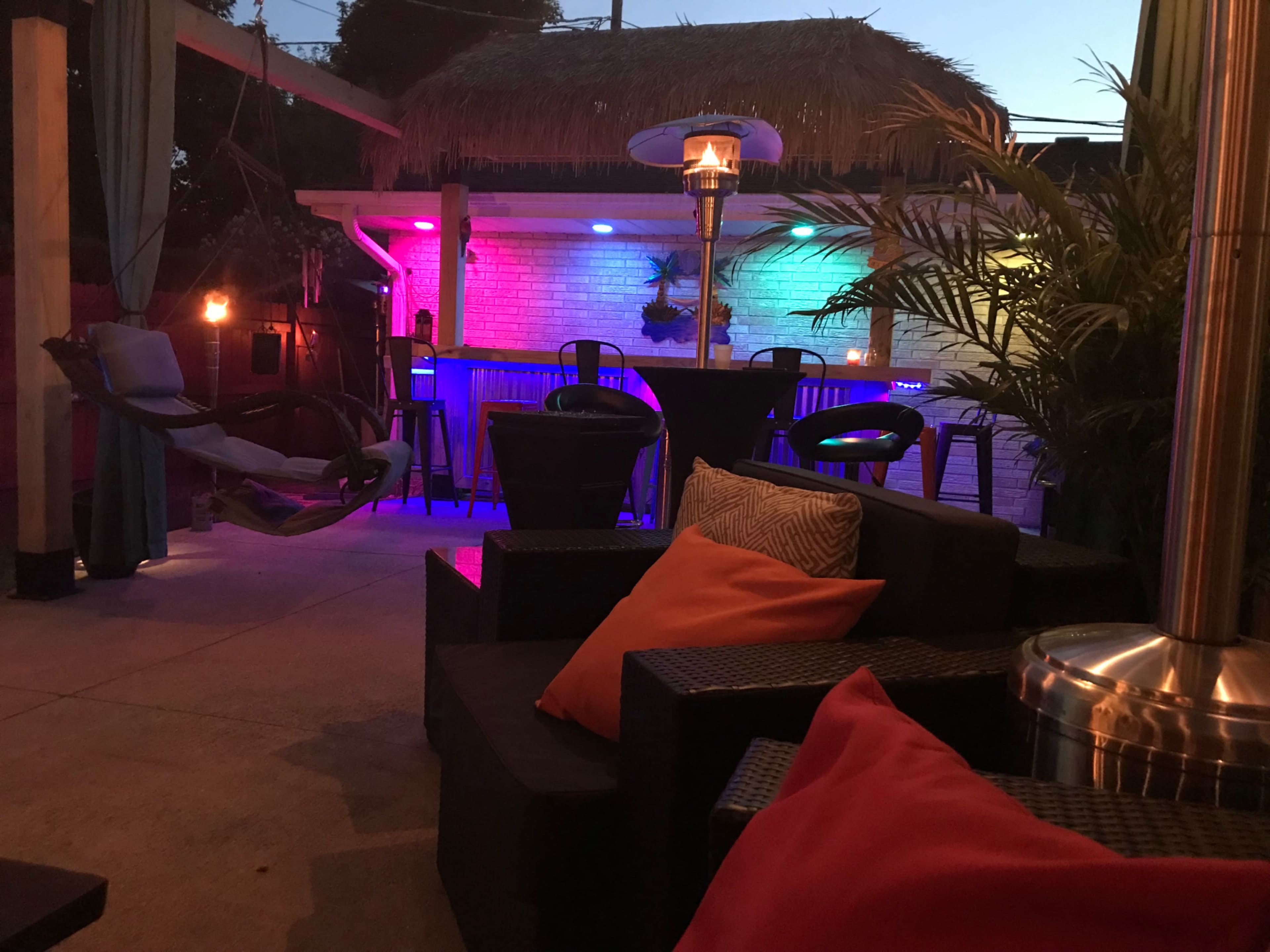 The scene depicts a cozy outdoor patio area with illuminated seating, a hammock, and a thatched roof bar, surrounded by palm plants.