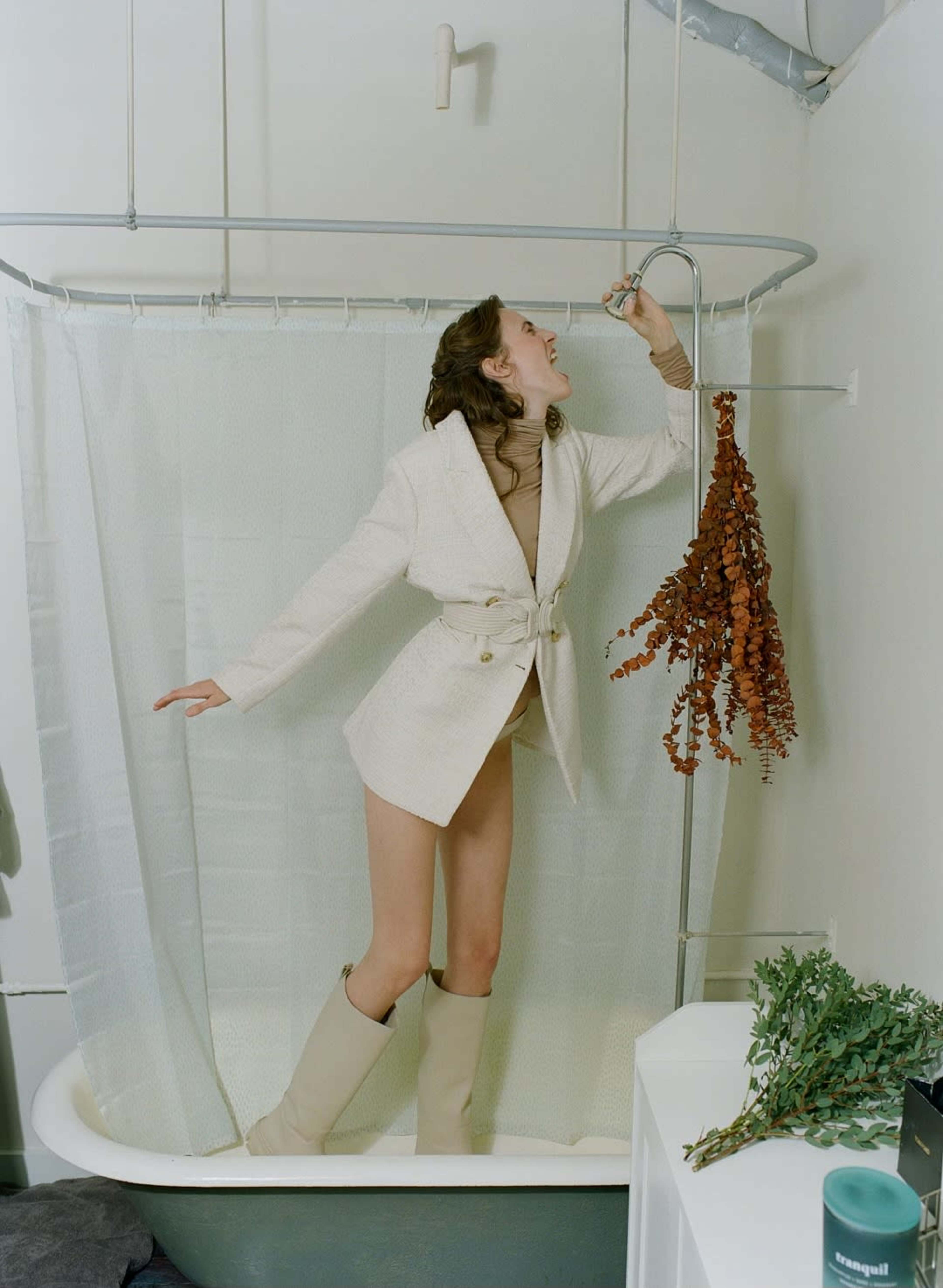 A person in a beige blazer and knee-high boots stands in a bathtub, reaching for a hanging showerhead while a cluster of dried plants decorates the side.