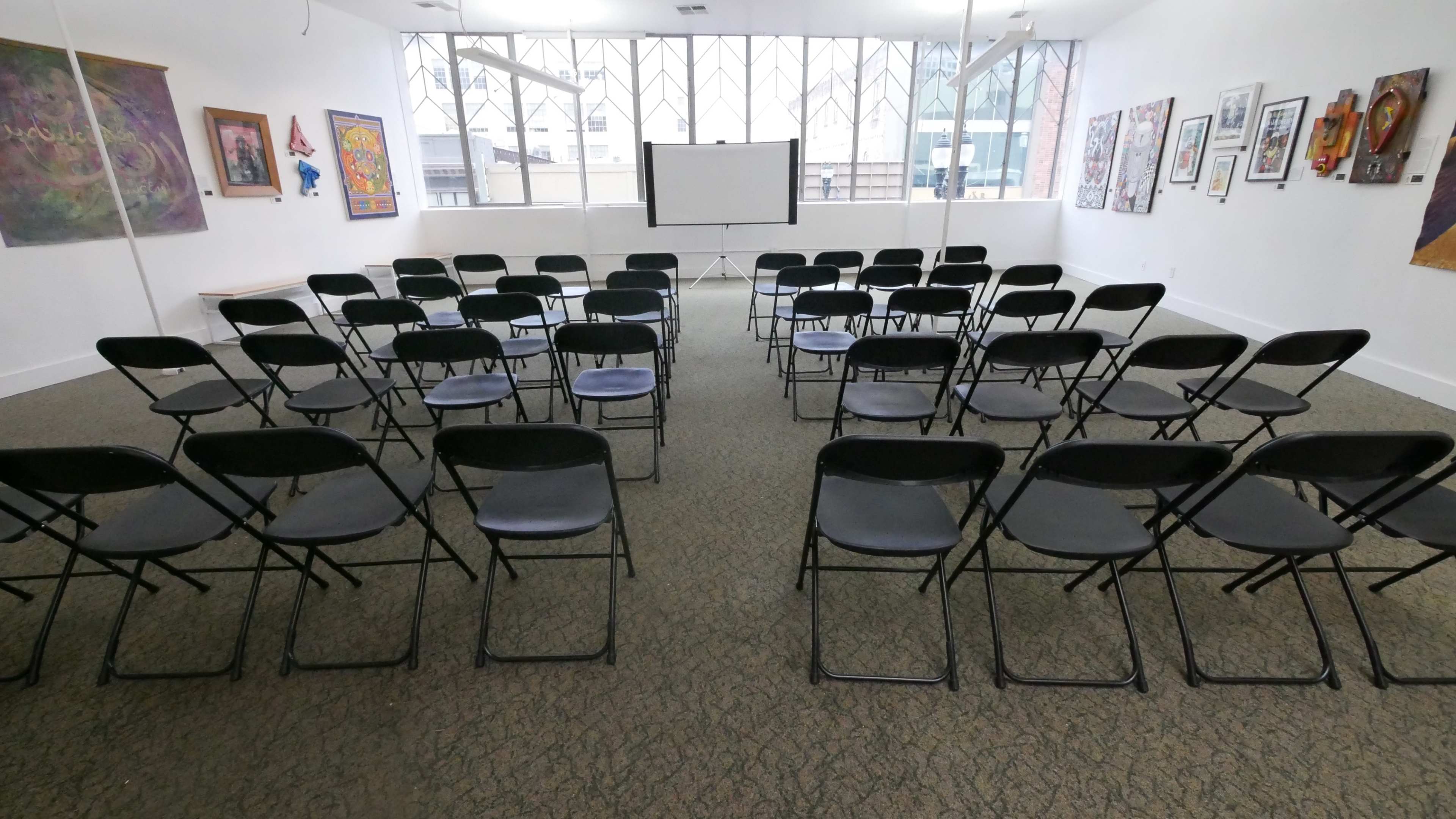 A room set up with rows of black folding chairs facing a white projection screen, surrounded by various framed artworks on the walls.
