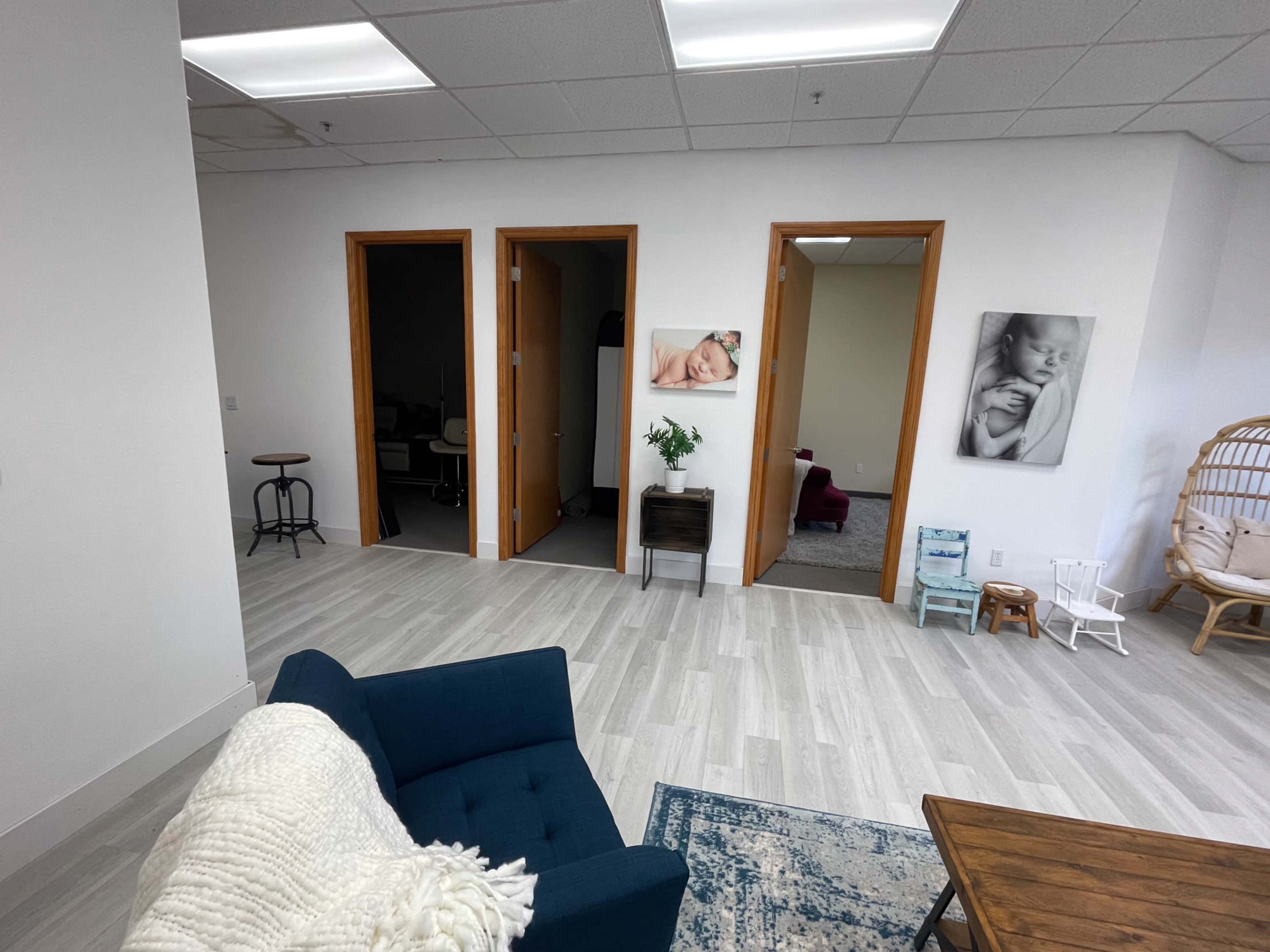 The image shows a modern office space featuring a blue couch, wooden table, and several doors leading to different rooms along with framed photographs on the wall.