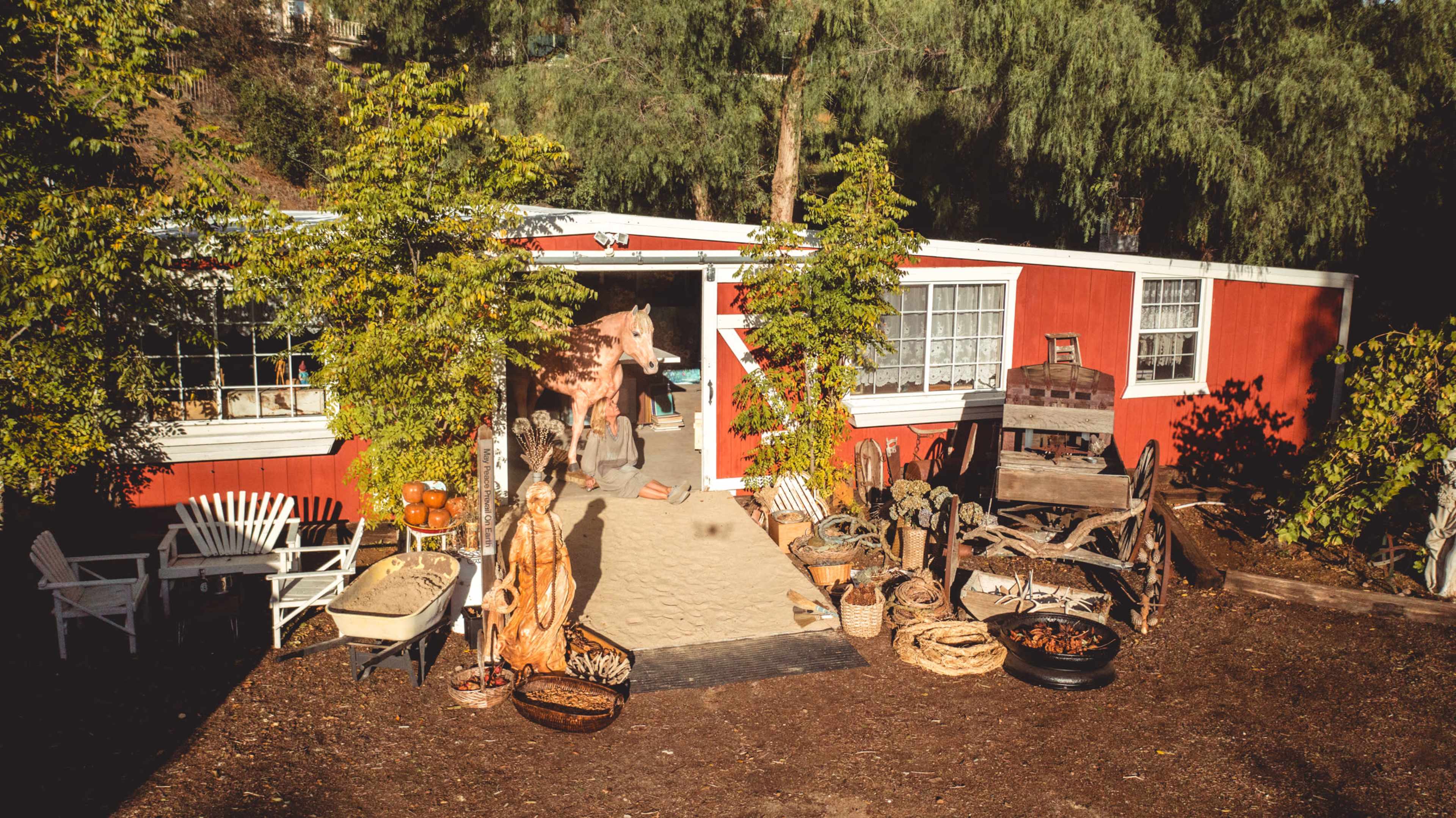 A red building with white trim features an entrance surrounded by various plants and rustic decor, including baskets, a wooden cart, and an array of sculptures.