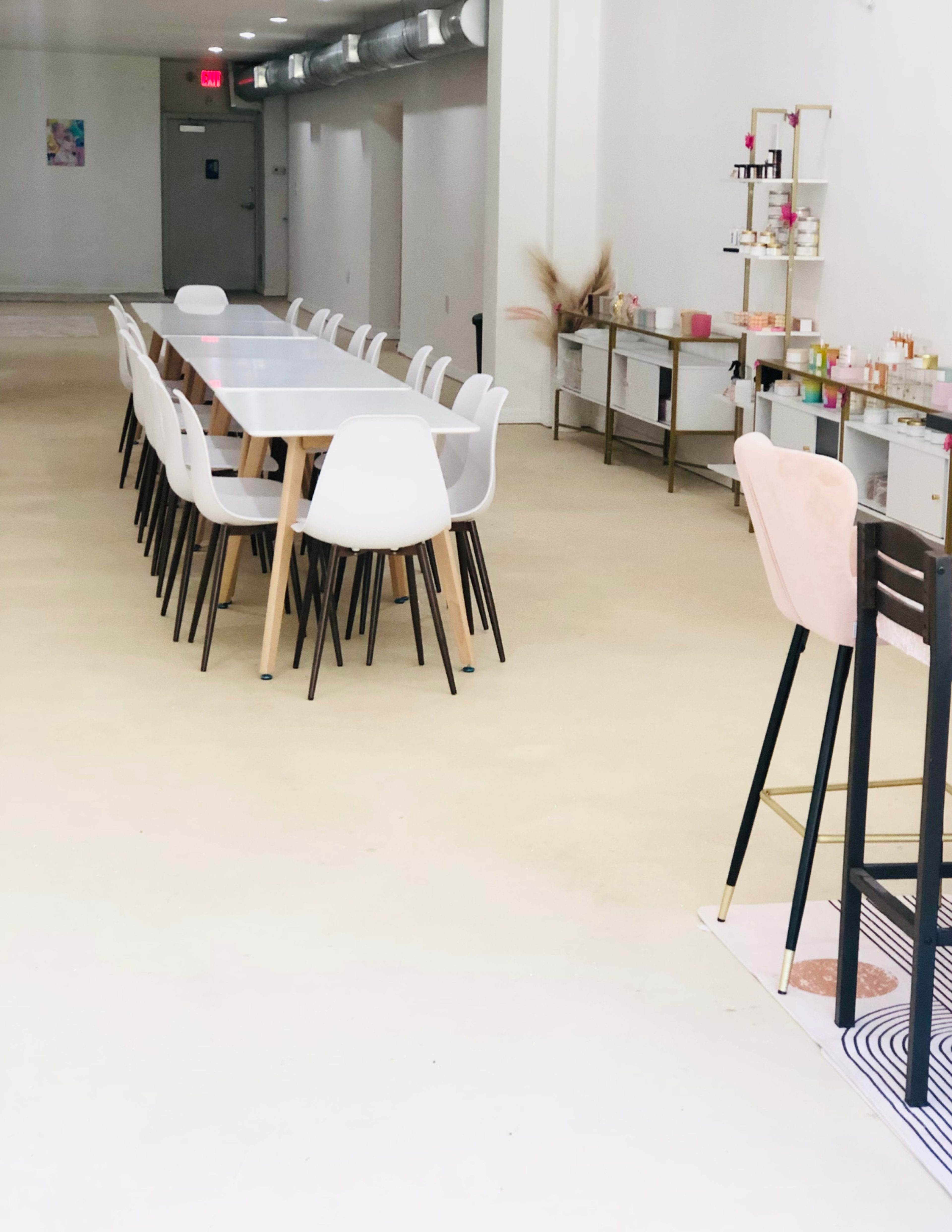 A long table with white chairs is set up in a spacious, minimalistic room featuring storage units and a bar area along one side.