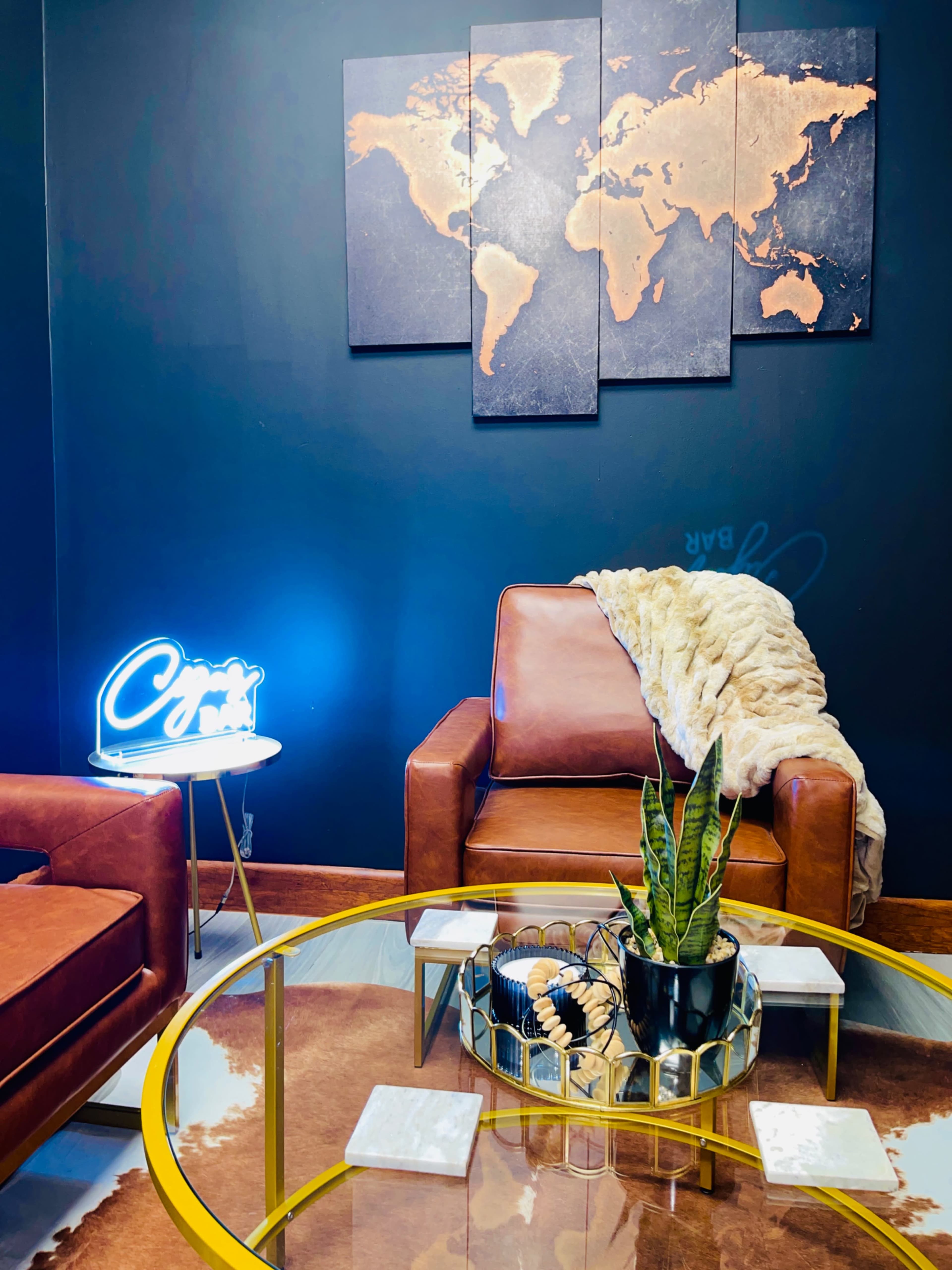 A cozy living area featuring two brown leather chairs, a round glass coffee table with decorative items, and a world map artwork on the wall, illuminated by a blue neon sign.