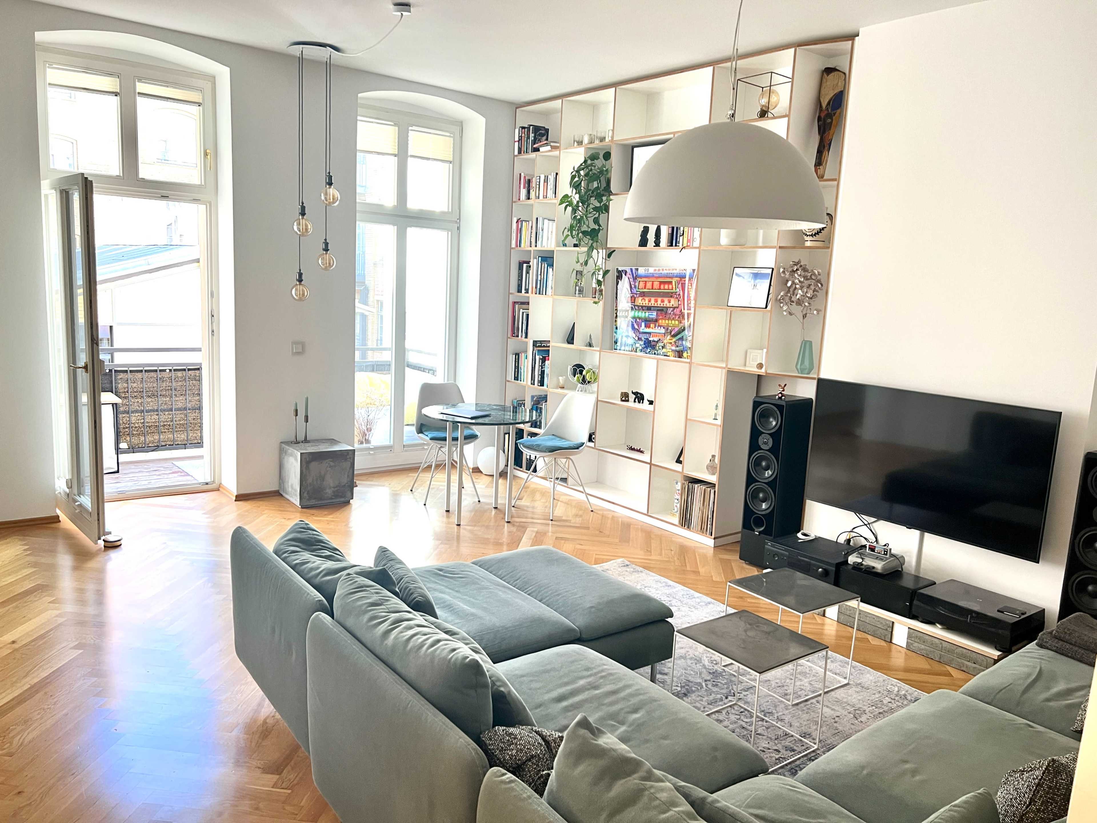 A modern living room featuring a light green sectional sofa, a glass coffee table, a television mounted on the wall, and a bookshelf filled with books, all with large windows allowing natural light.