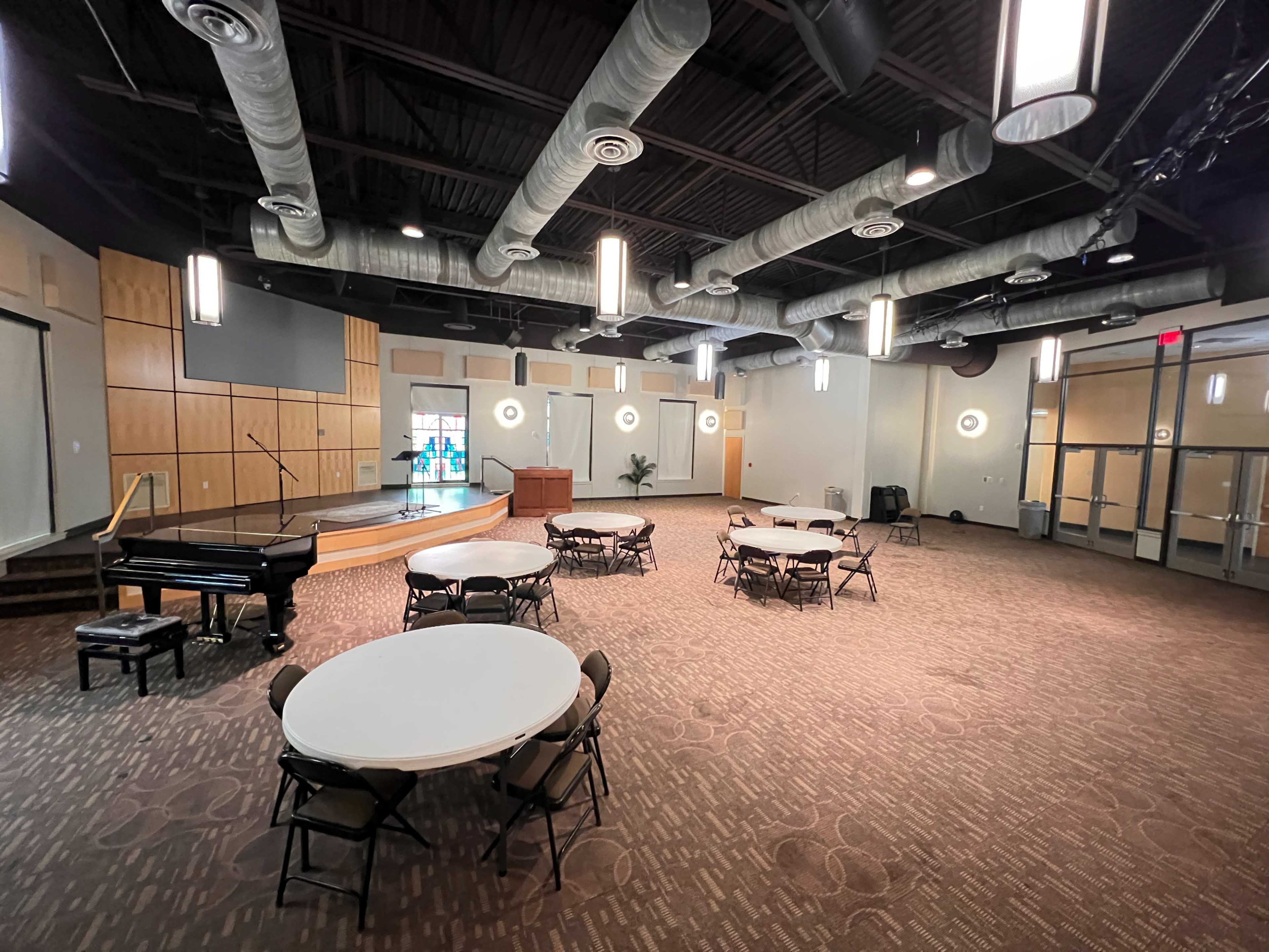 The image shows a spacious indoor venue featuring several round tables set up, a grand piano in the corner, and a stage area at the front.