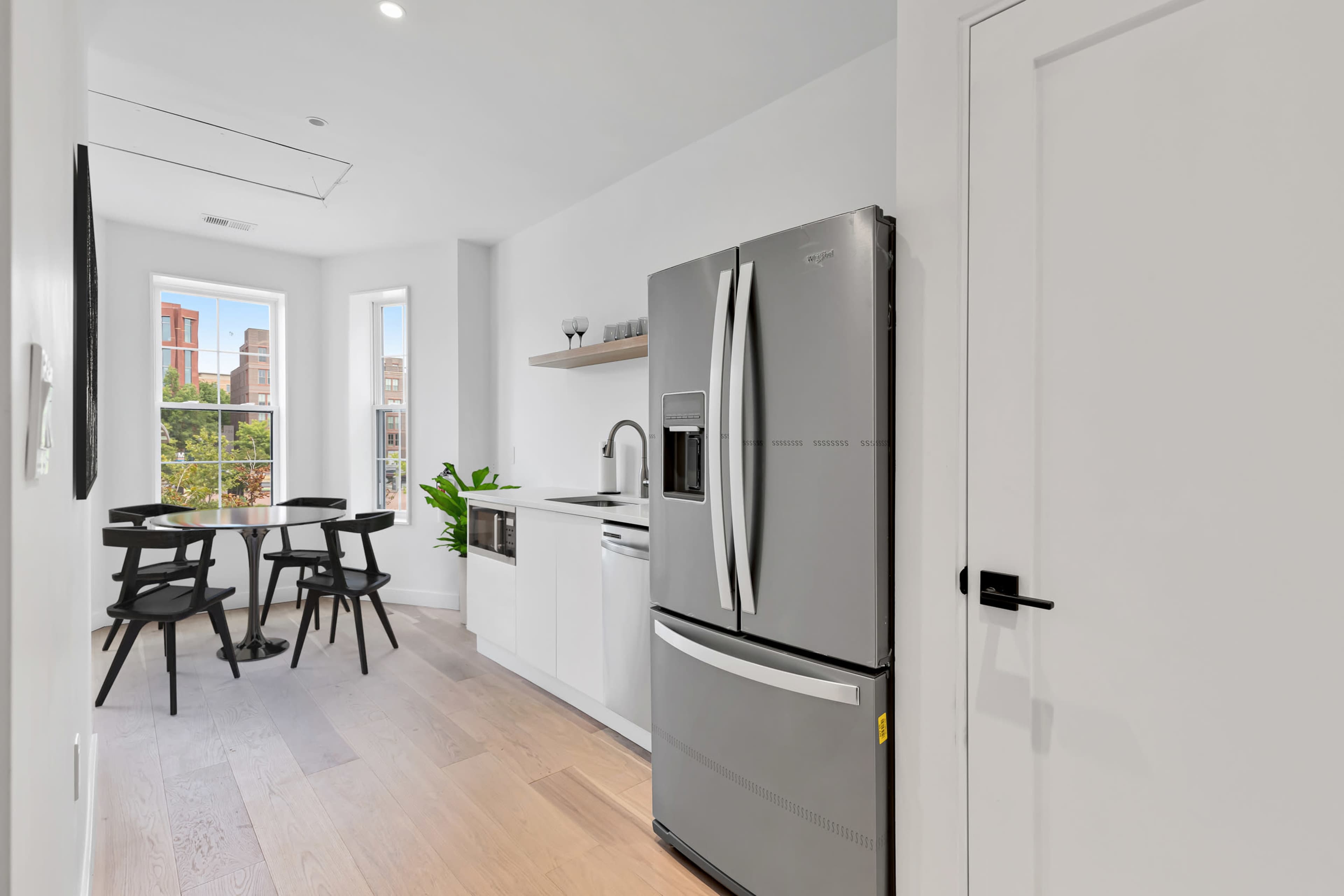 A modern kitchen features a refrigerator, sink, and a small dining table with four black chairs beside large windows.