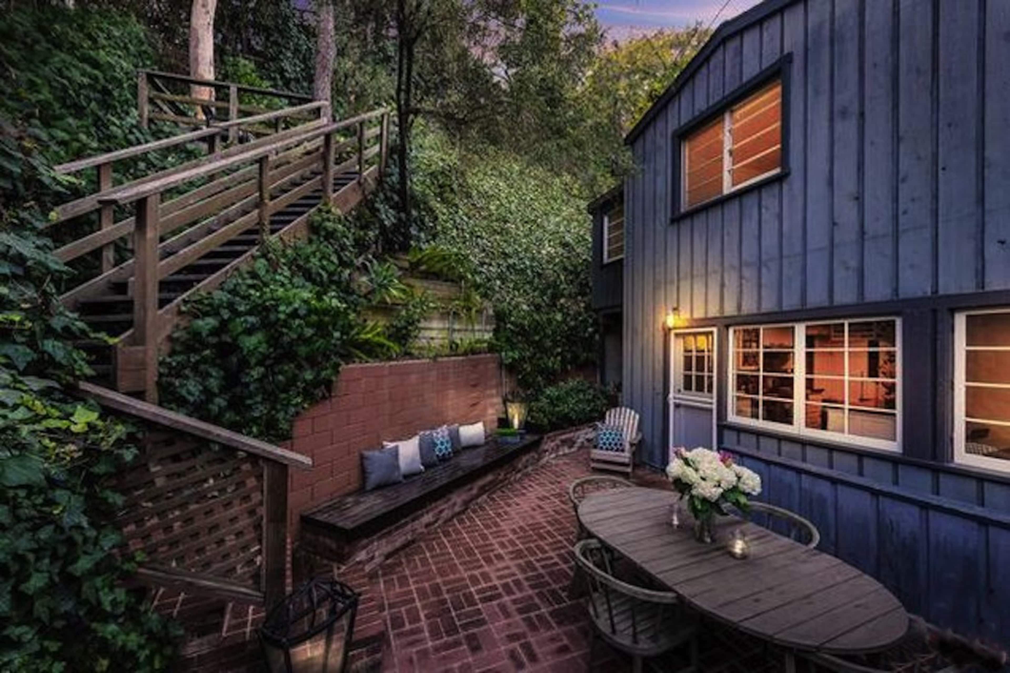 A blue wooden house is nestled among greenery, featuring a brick patio with outdoor seating and a staircase leading up to a higher level.