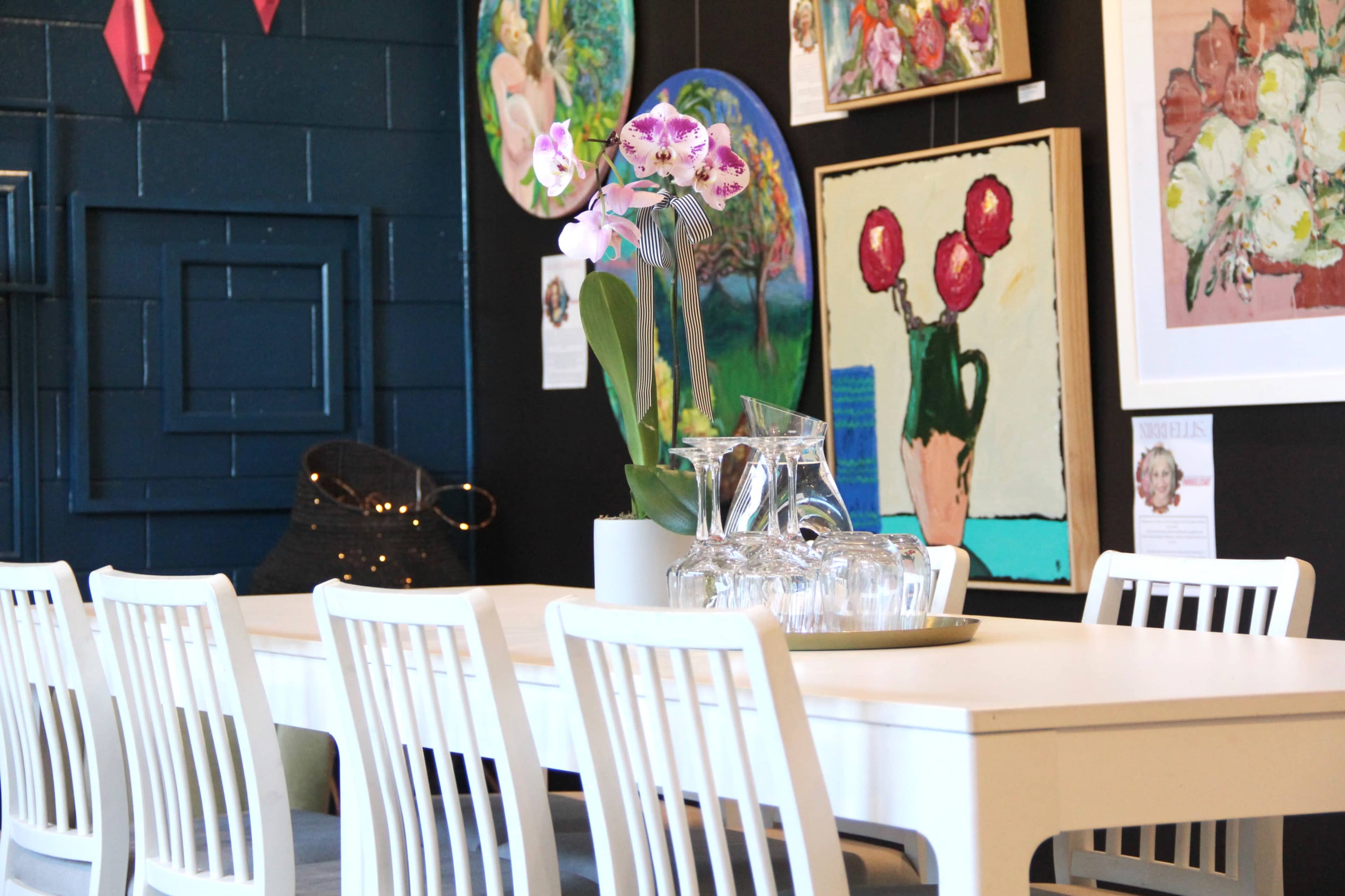 A white table is set with a pitcher and glasses, surrounded by chairs, in a room displaying colorful artwork on the walls.