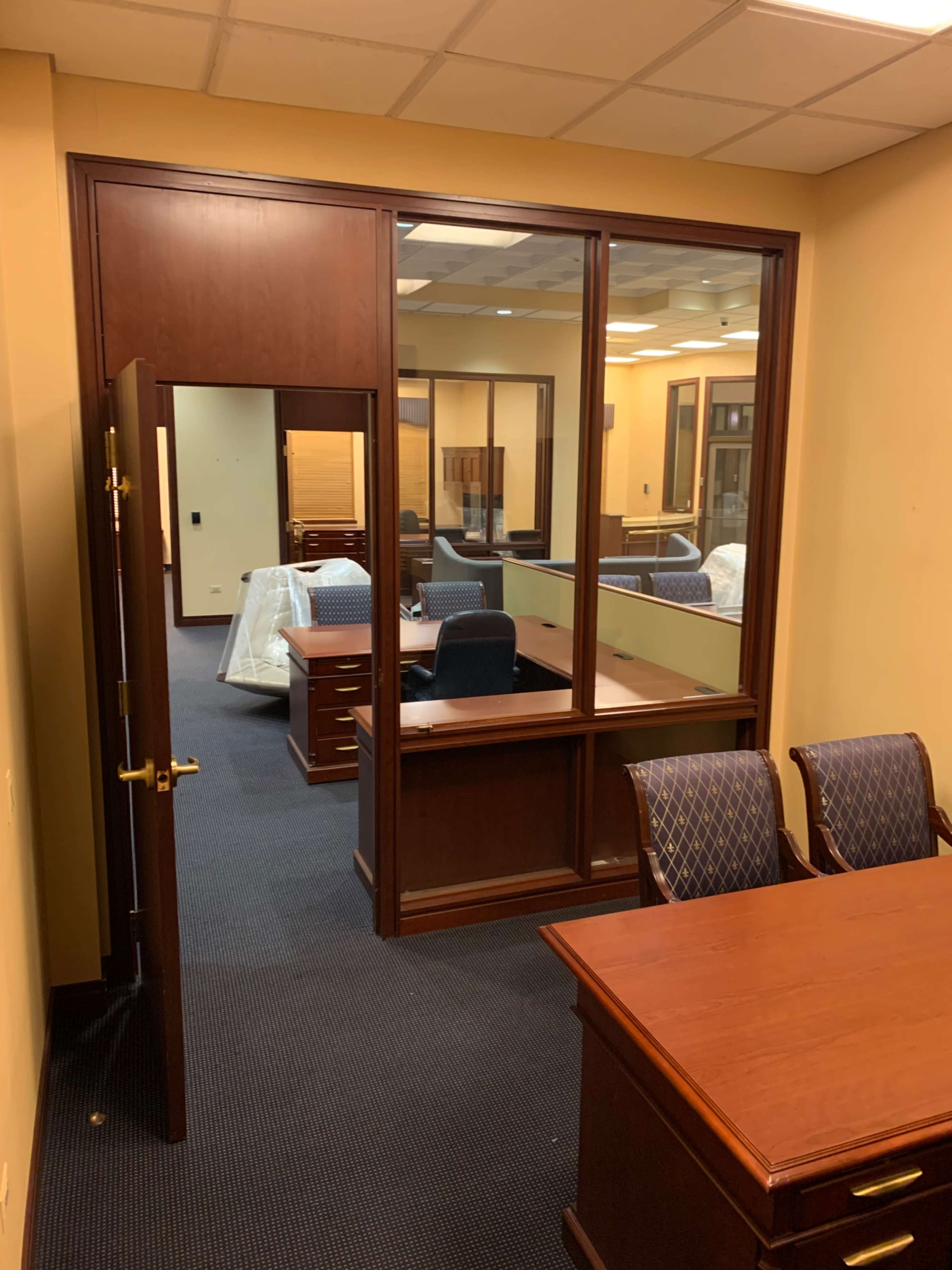 An office space features a wooden desk, two chairs, and a glass-paneled partition with an open doorway leading to another area filled with additional desks and chairs.