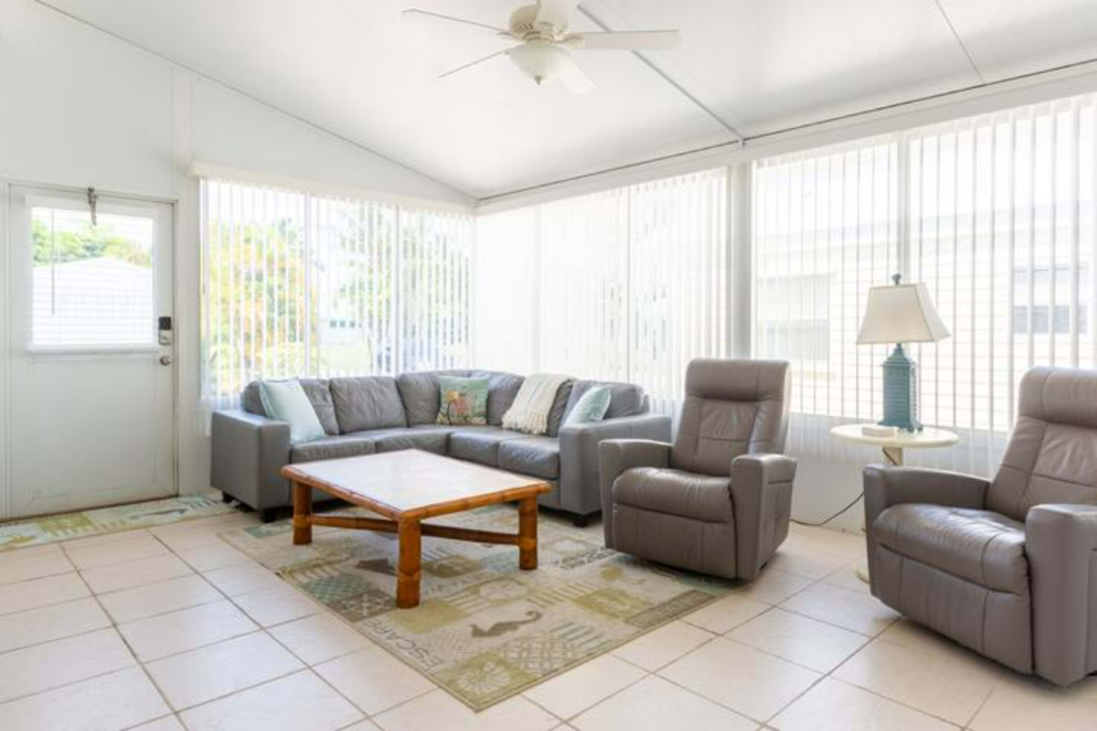 A sunlit sunroom featuring a gray sectional sofa, two reclining chairs, a wooden coffee table, and a lamp beside the windowed walls.
