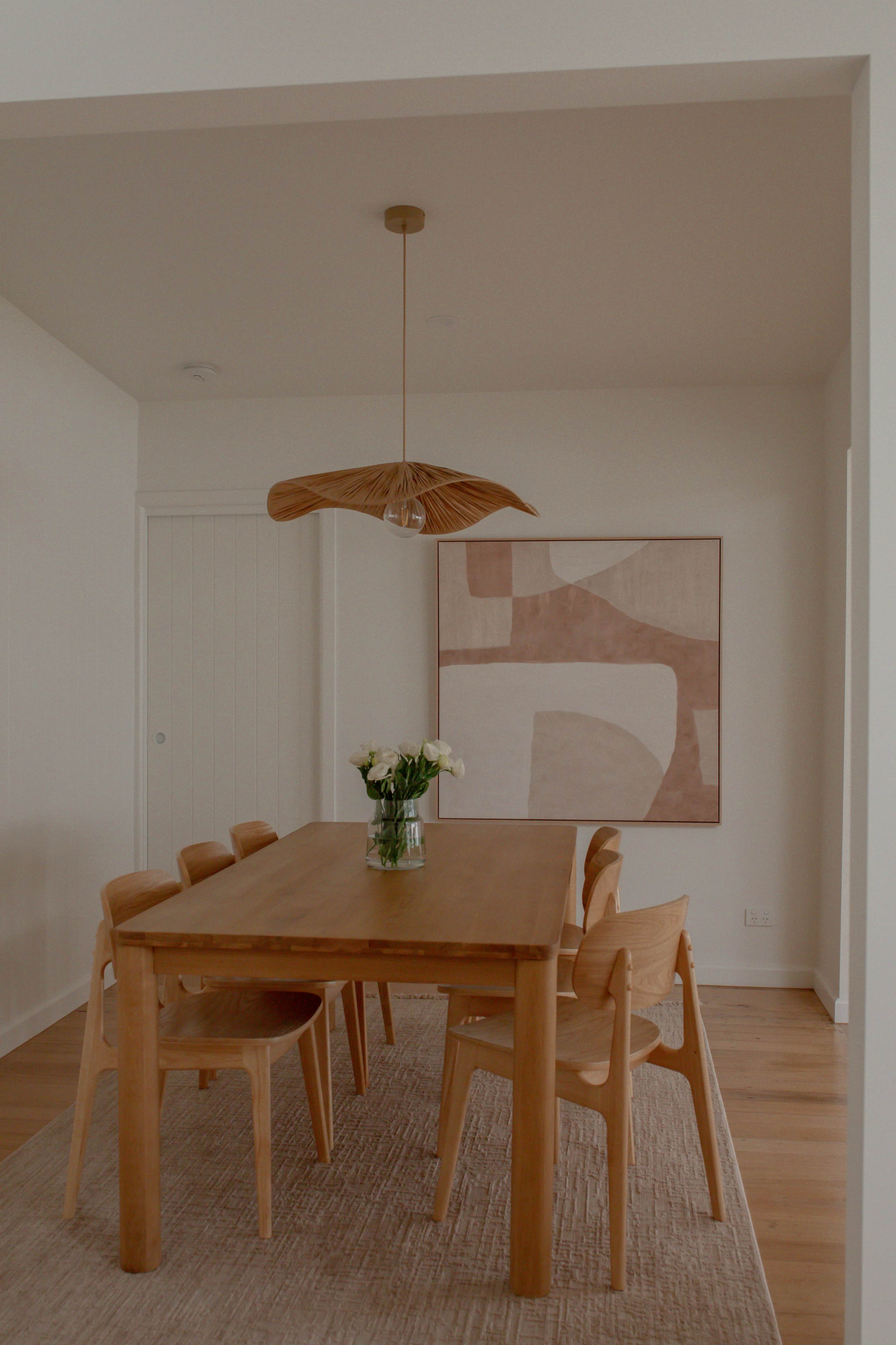A wooden dining table with four chairs is set in a room featuring a large abstract artwork on the wall and a pendant light hanging above.