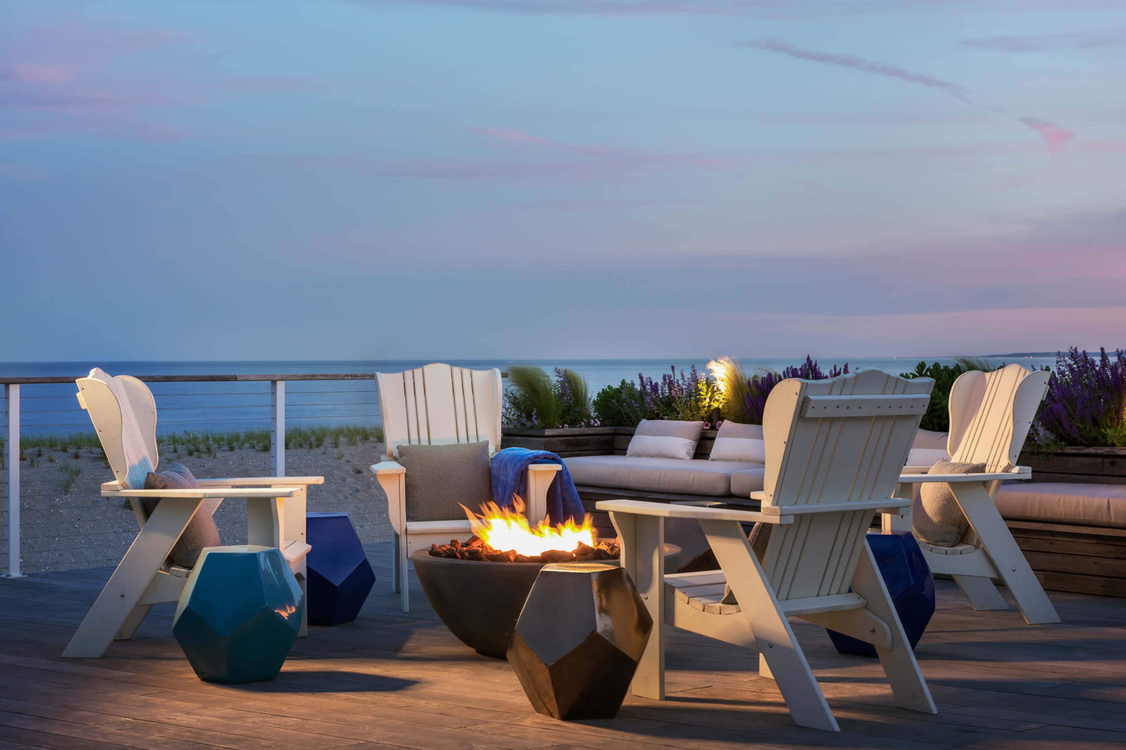 A cozy outdoor seating area features four white Adirondack chairs surrounding a fire pit on a deck overlooking the beach at dusk.