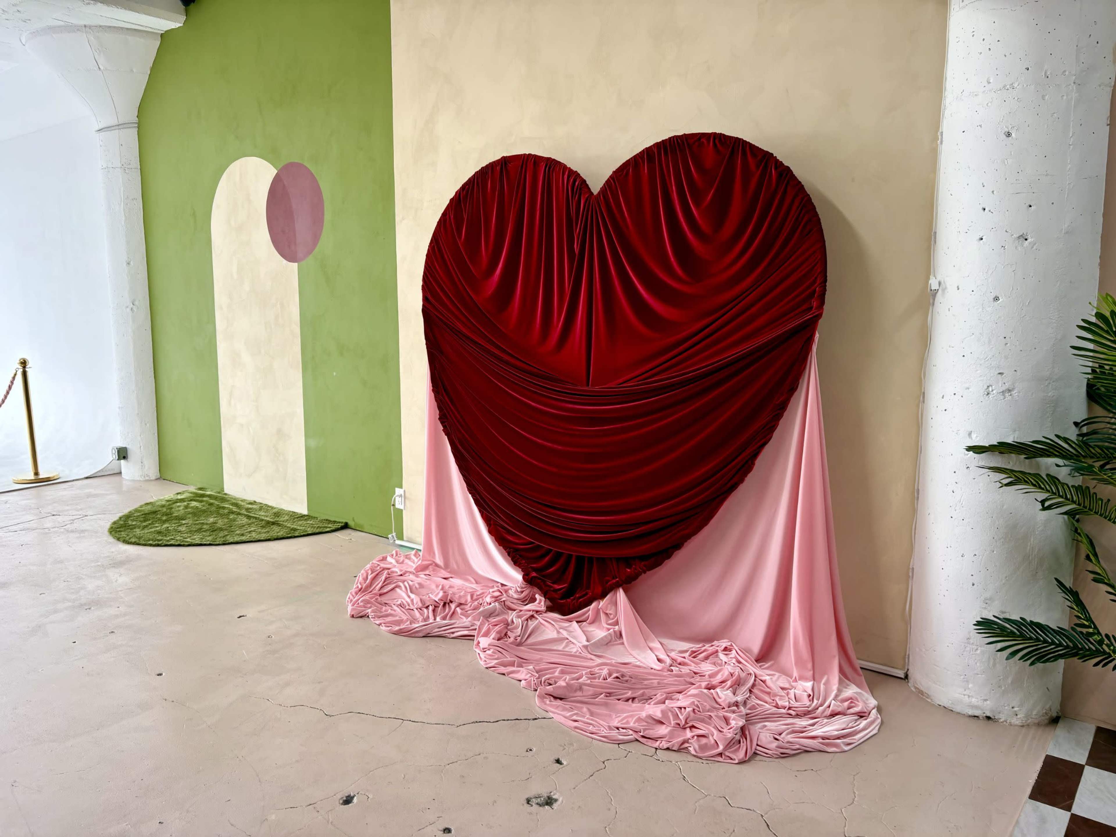 A large, red heart-shaped fabric installation drapes over the floor against a light-colored wall, complemented by pink fabric below it.