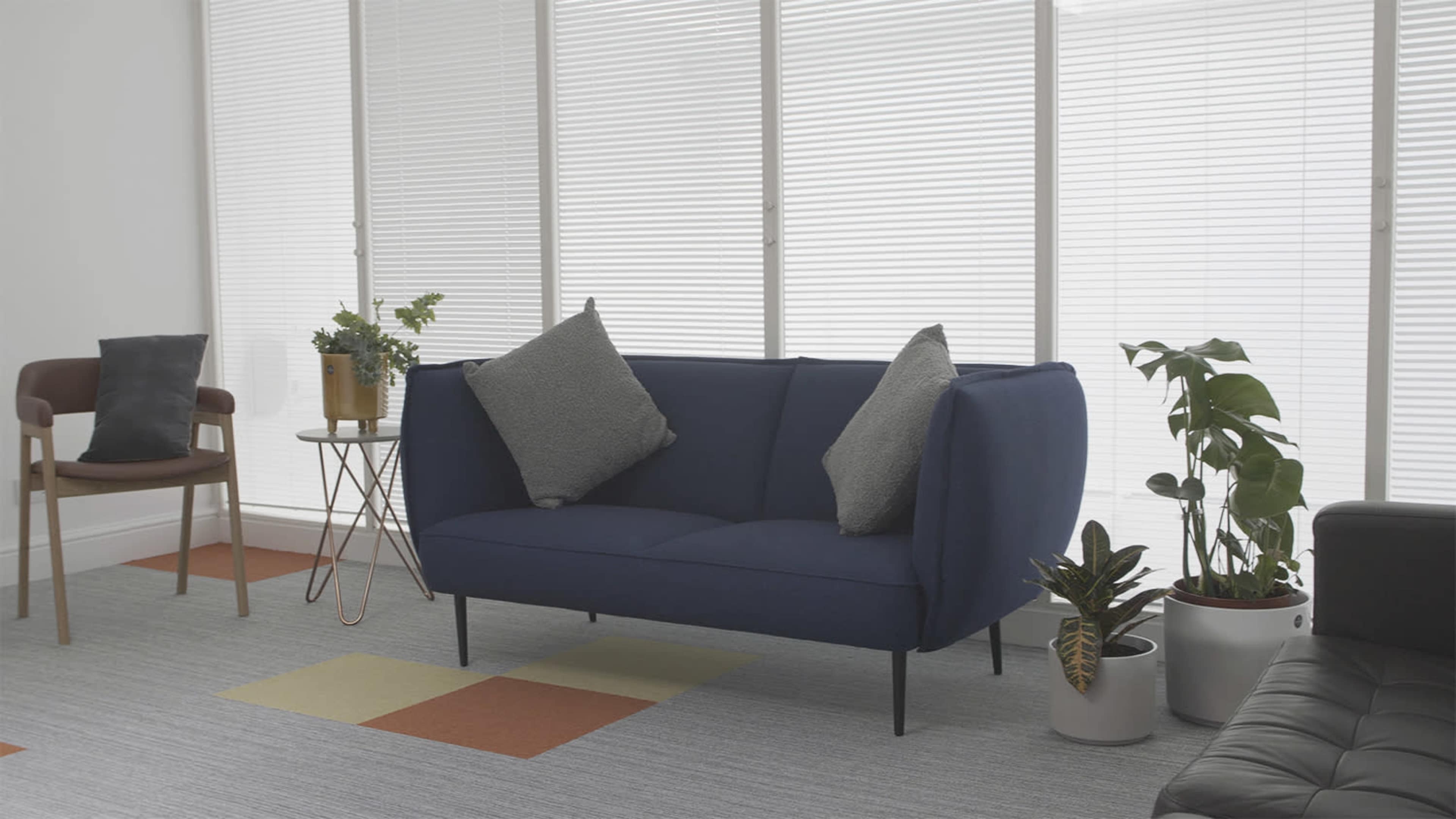 A modern living space featuring a blue sofa with two cushions, a small round table, a chair, and several potted plants arranged near large windows with blinds.