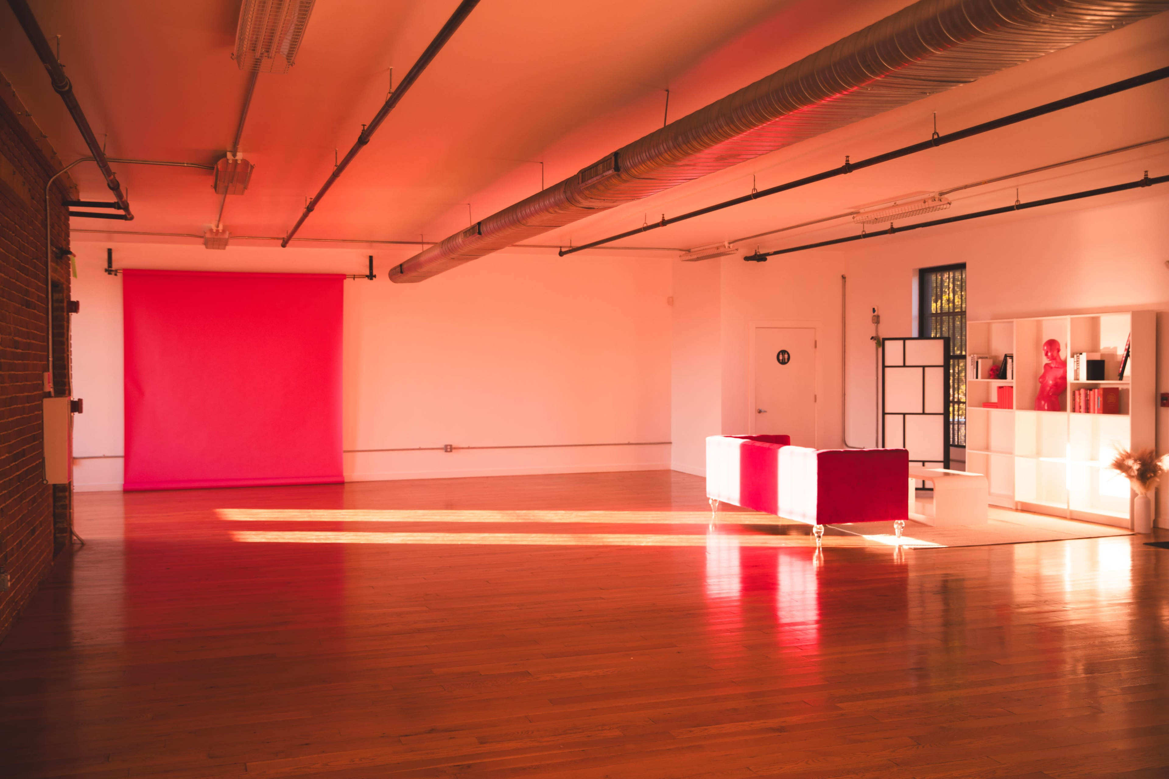 A spacious room features wooden flooring, exposed brick walls, and a bright pink backdrop along with a red sofa and a white shelving unit.
