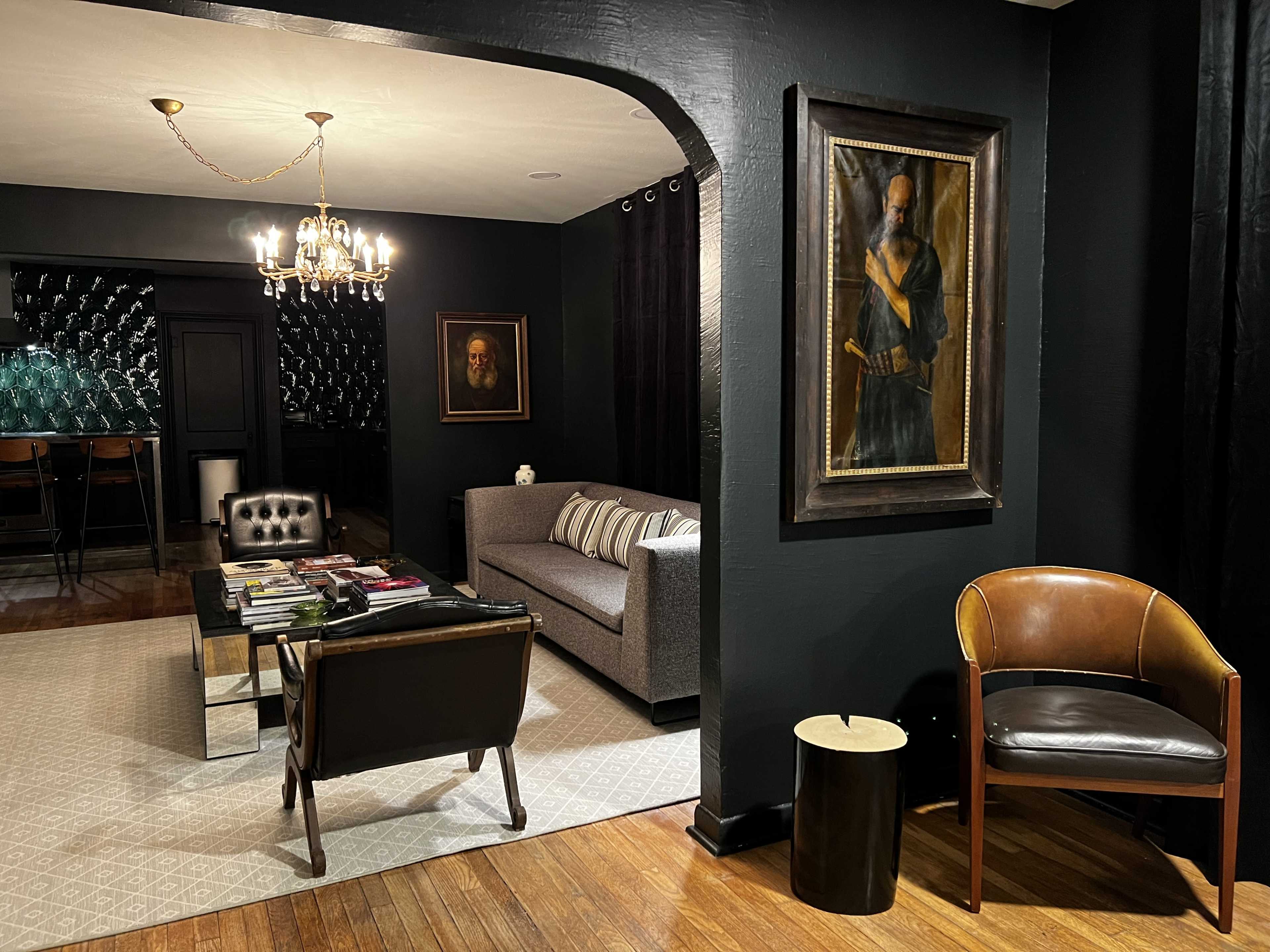 A stylish living room featuring a gray sofa, a black leather chair, a wooden coffee table with magazines, and two framed portraits on the wall, all set against dark-colored walls.