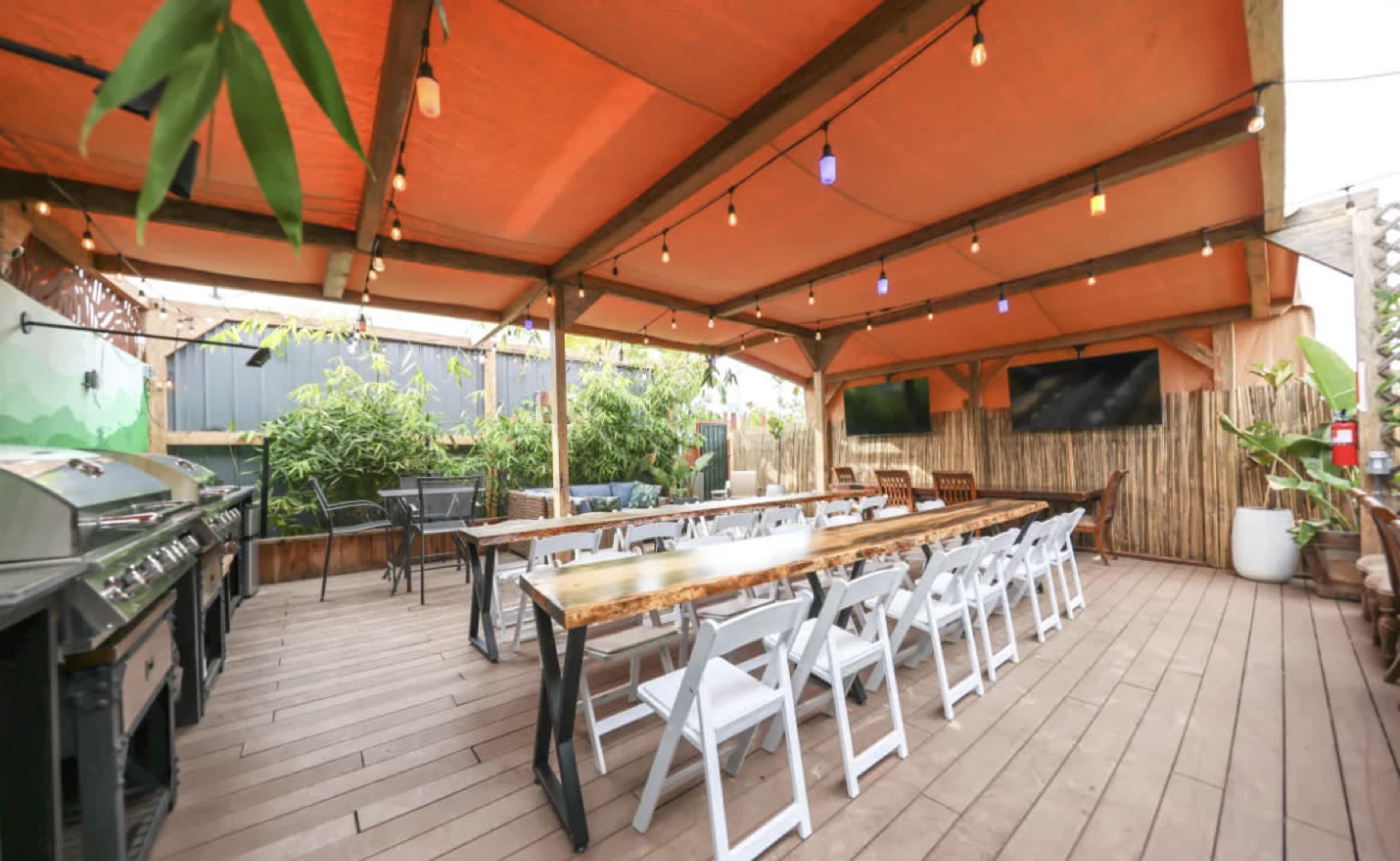 The image shows an outdoor dining area with a long wooden table, white folding chairs, string lights, and a grill under a large orange canopy.