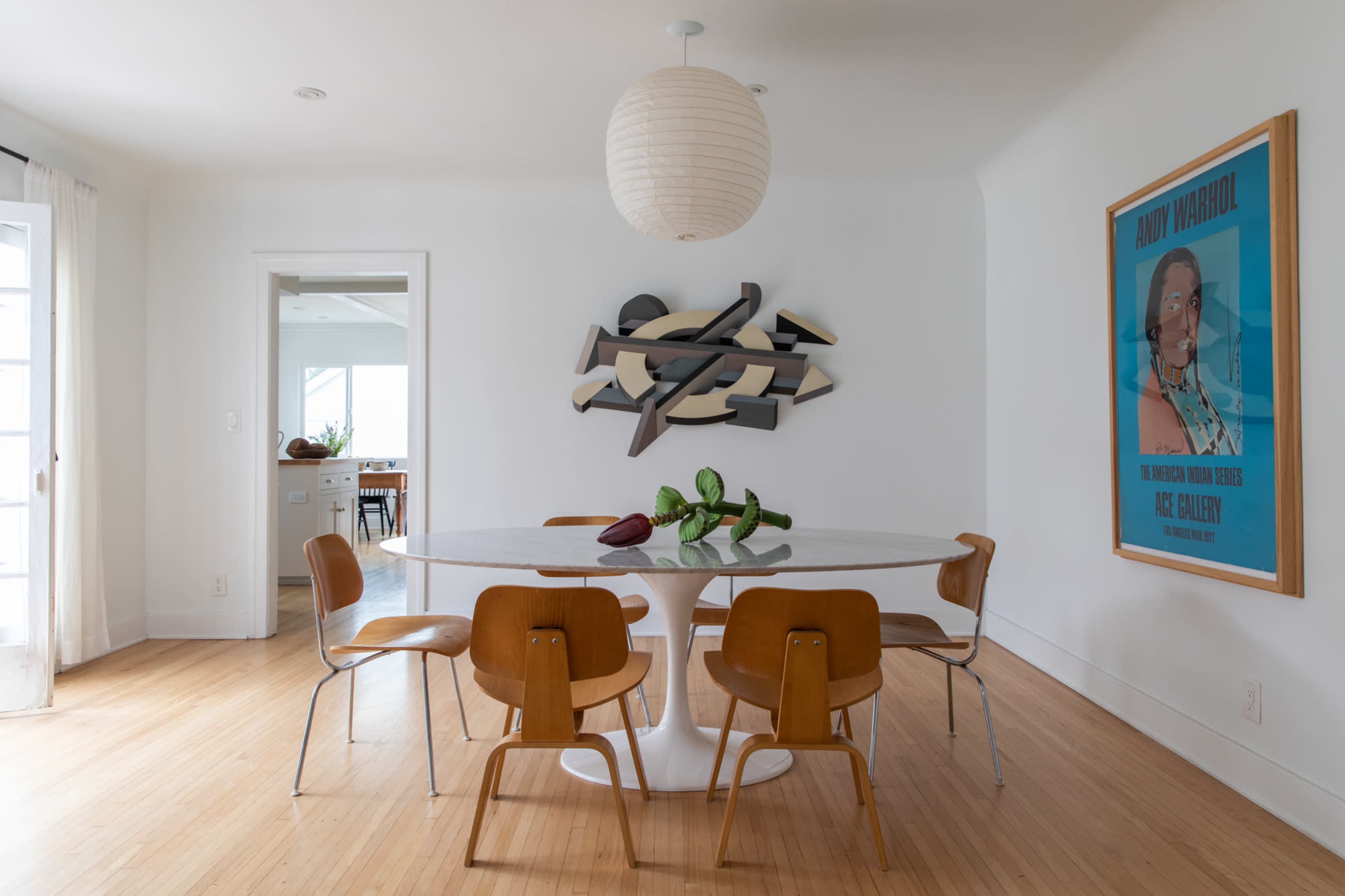 A dining area features a round table surrounded by four wooden chairs, with a large artwork on the wall opposite a door leading outside.