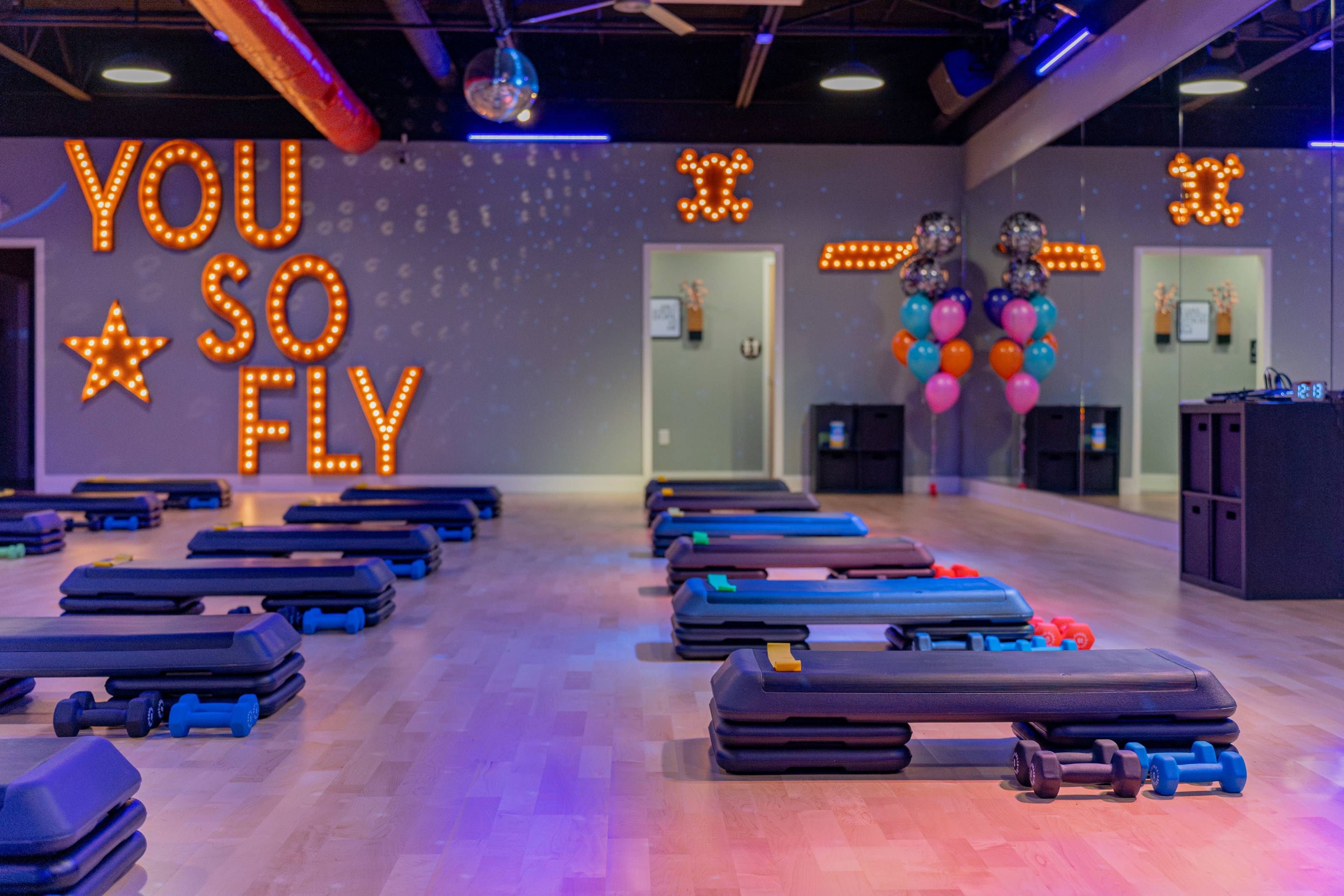 A fitness studio is set up with exercise mats and weights, featuring a brightly lit sign that reads "YOU SO FLY" on one wall.