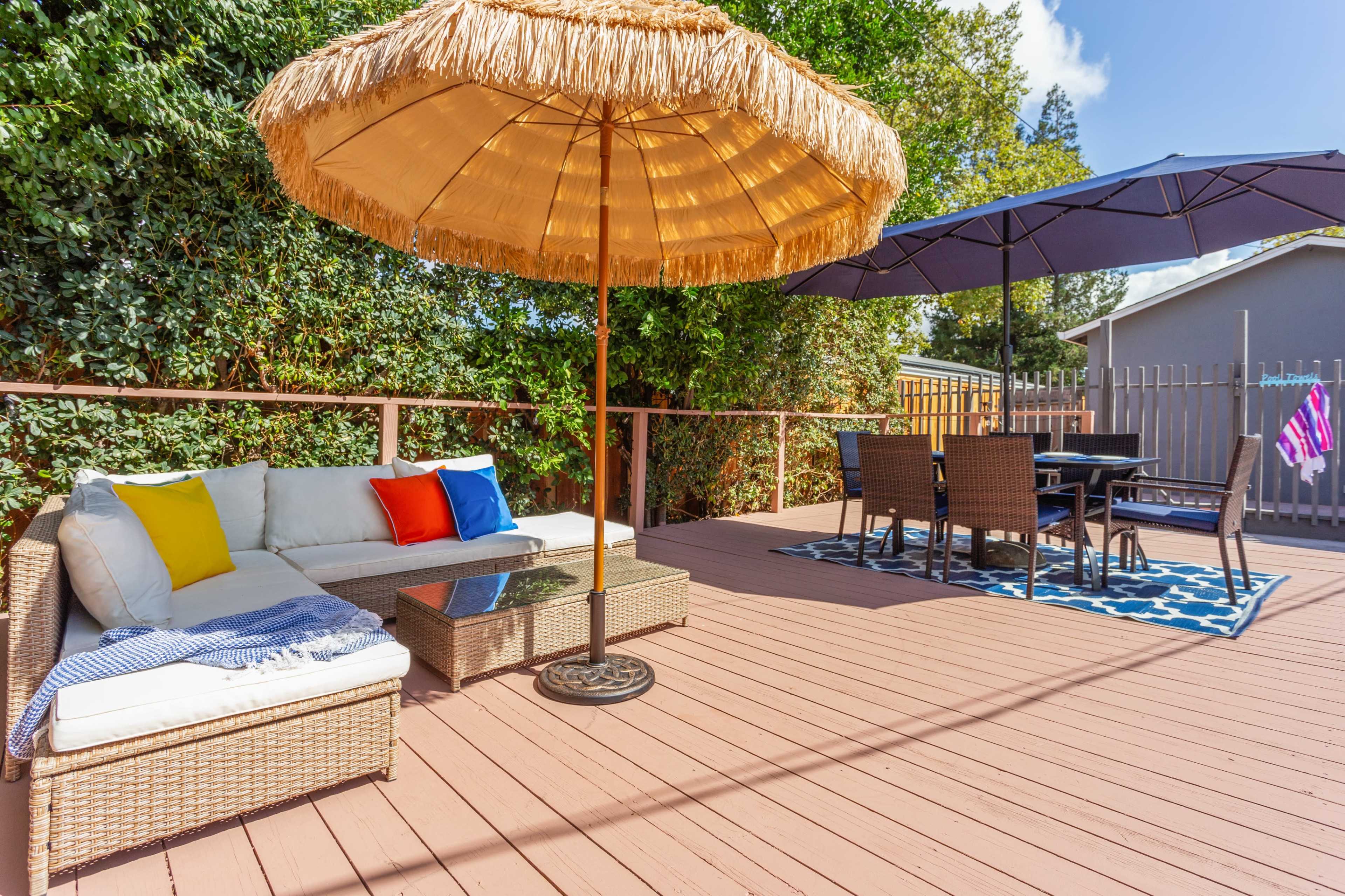 A patio features a wicker seating area with colorful pillows, a thatched umbrella, and a dining set arranged on a patterned rug.