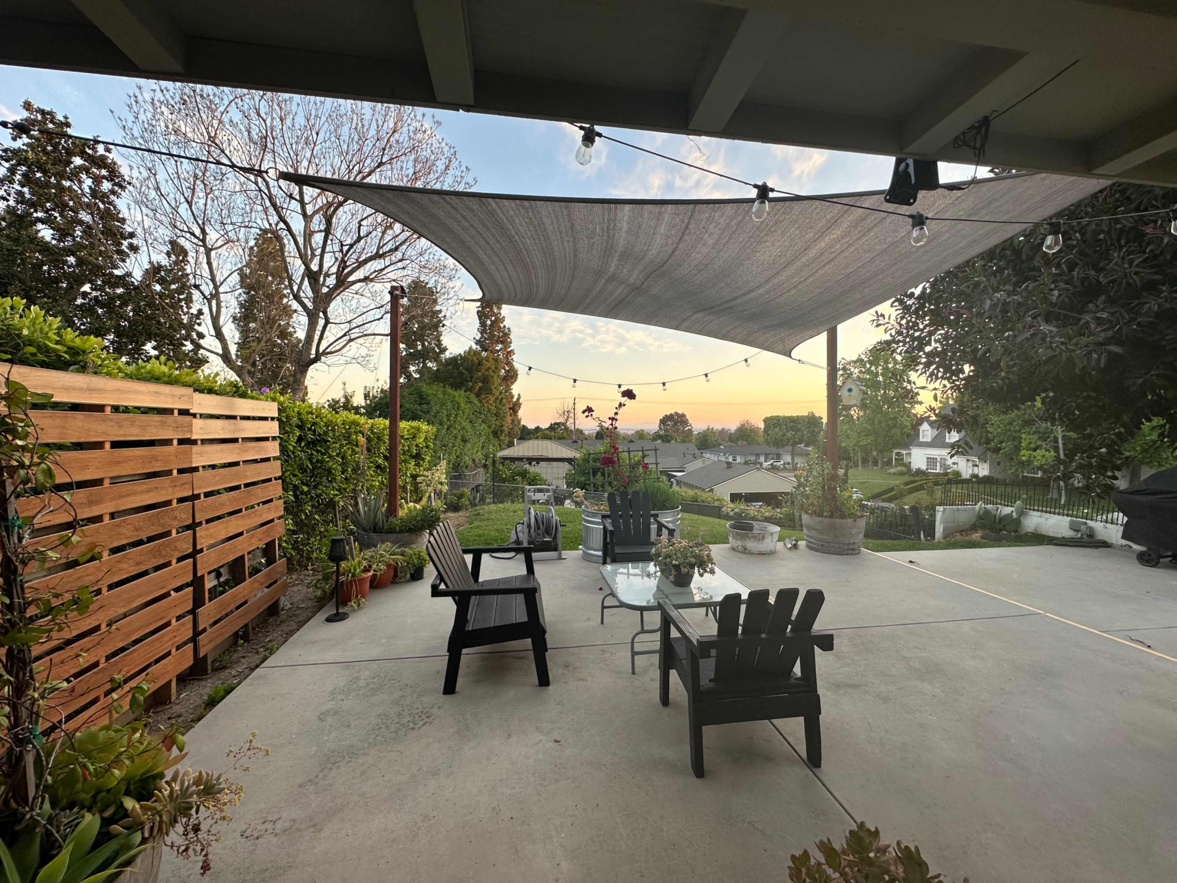 A patio with modern furniture, a shade sail overhead, and potted plants, overlooking a suburban area at sunset.