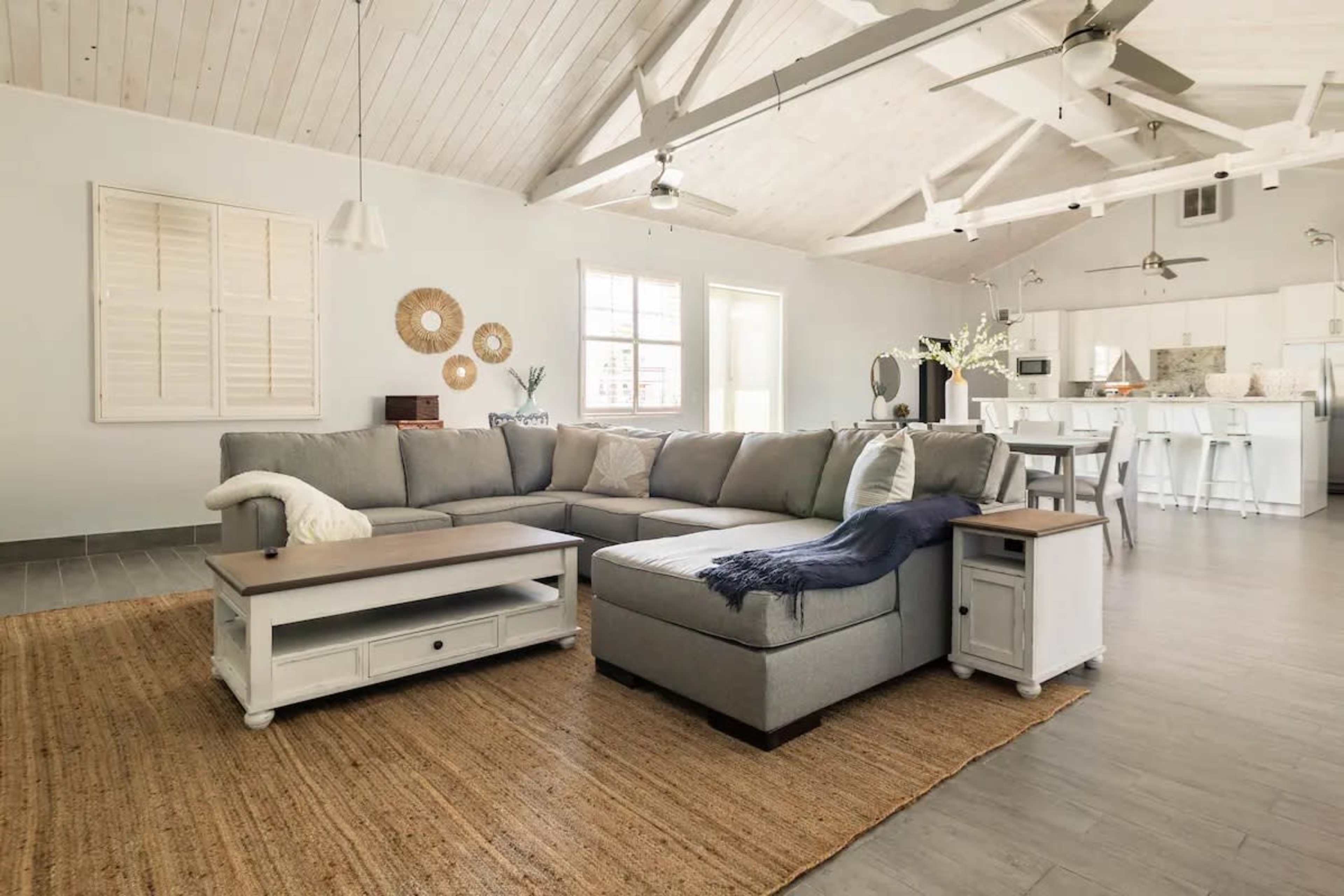 A spacious living room features a gray sectional sofa, a light wooden table, and a woven area rug beneath a vaulted ceiling.