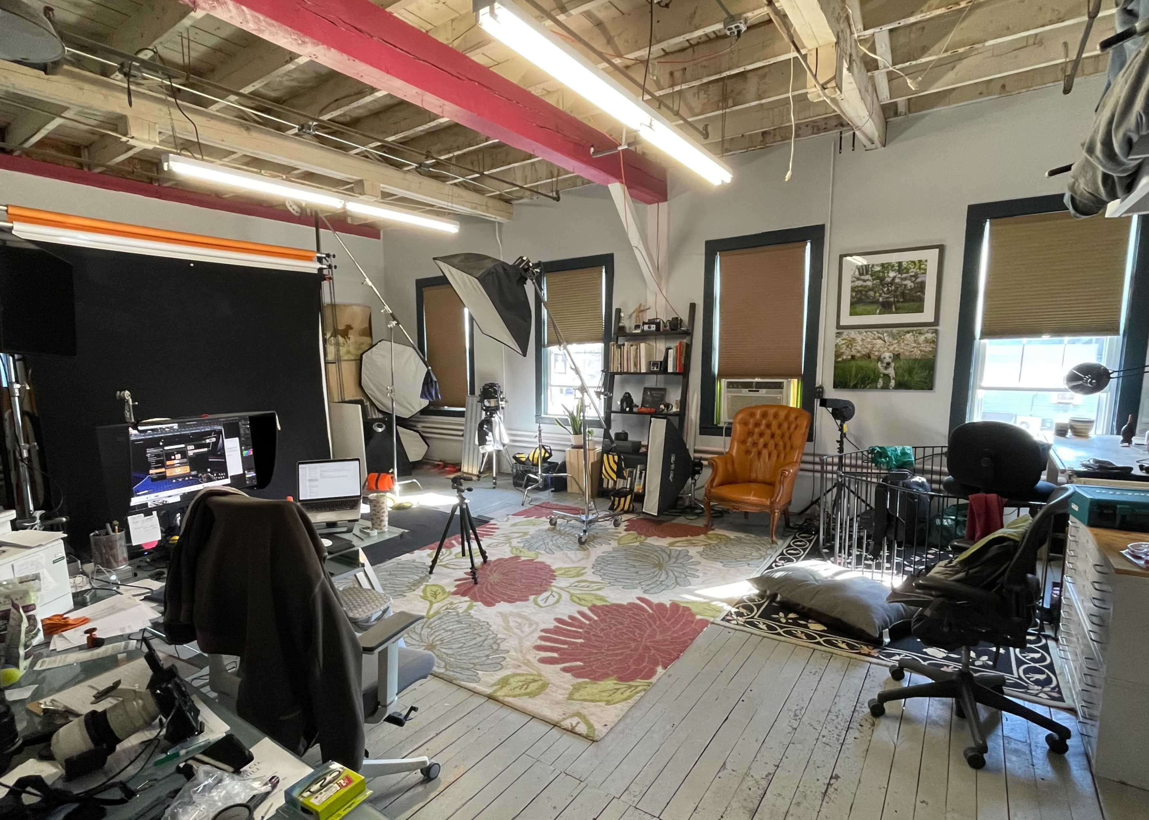 A cluttered studio space with various photography equipment, a large area rug, multiple chairs, and a computer workstation.