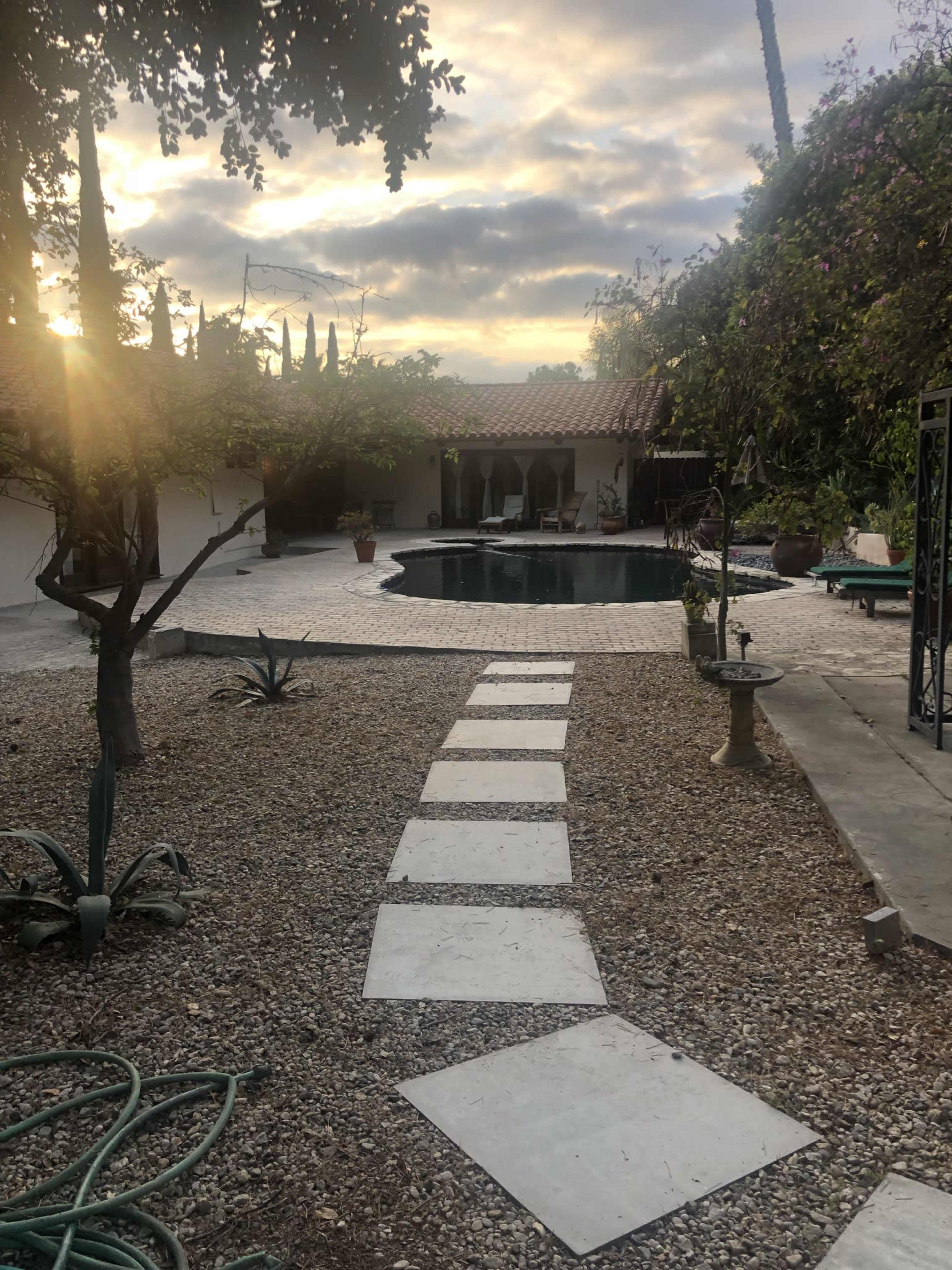A stone pathway leads to a swimming pool surrounded by greenery and a patio under a cloudy sky.