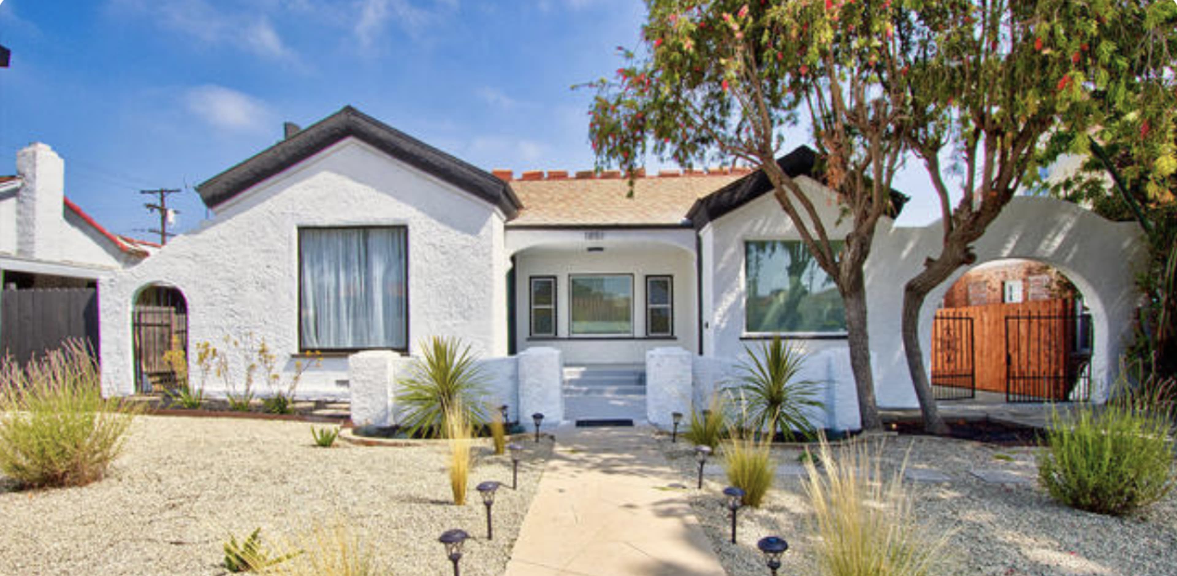 The image shows a single-story white house with a red-tiled roof, surrounded by a landscaped gravel yard and ornamental plants.