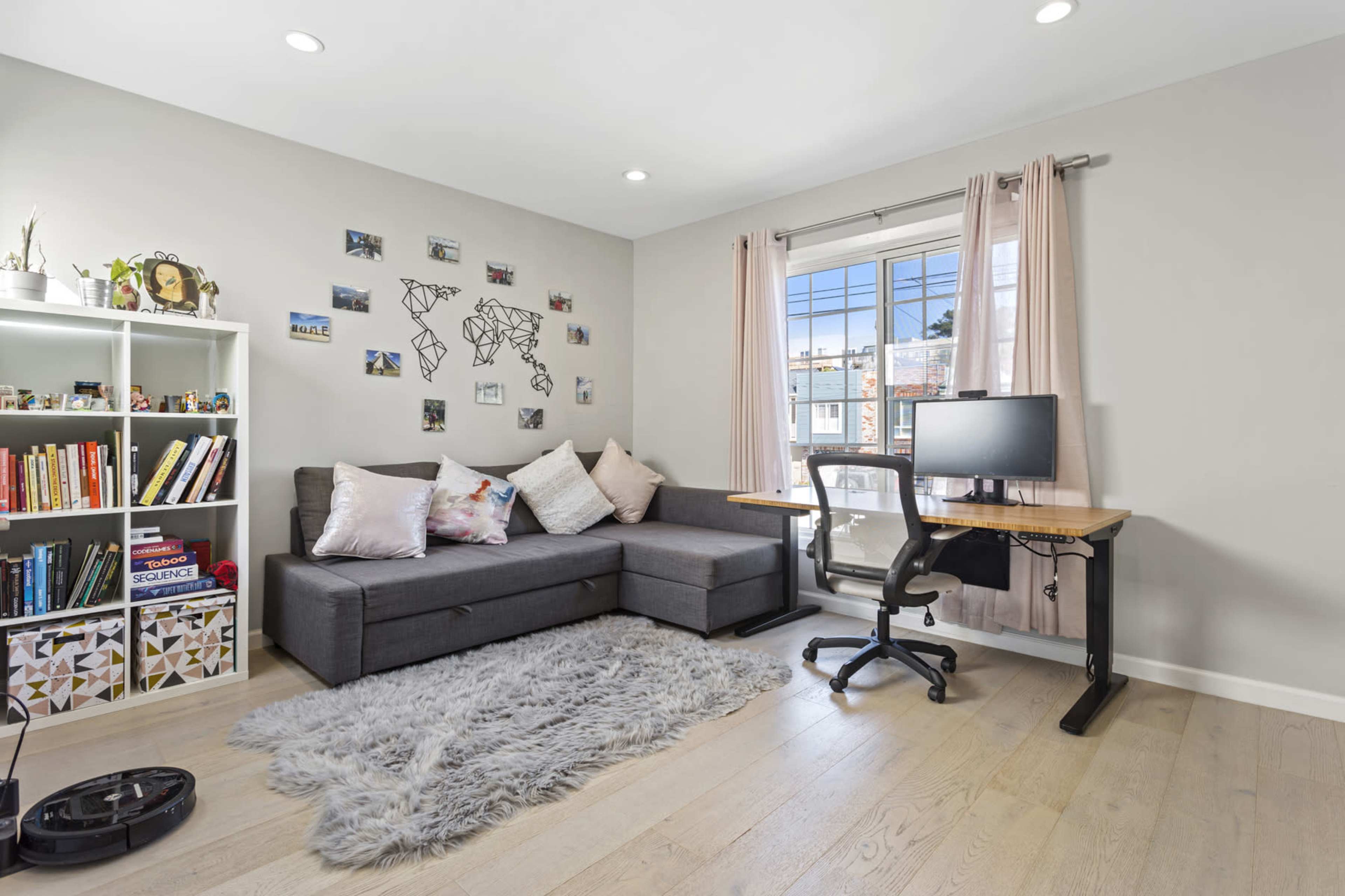 A modern home office with a sectional sofa, a wall decorated with photos and a map, a desk with a computer, and a window allowing natural light to enter.