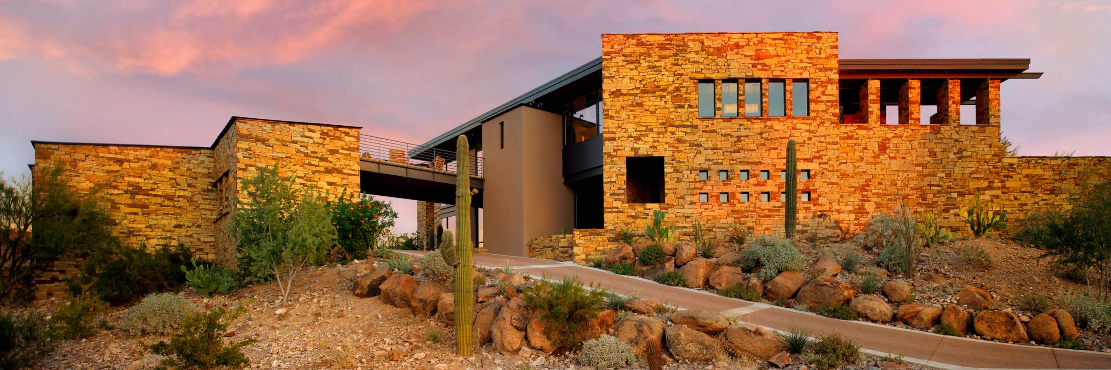 A modern stone house with large windows, set against a desert landscape at sunset.