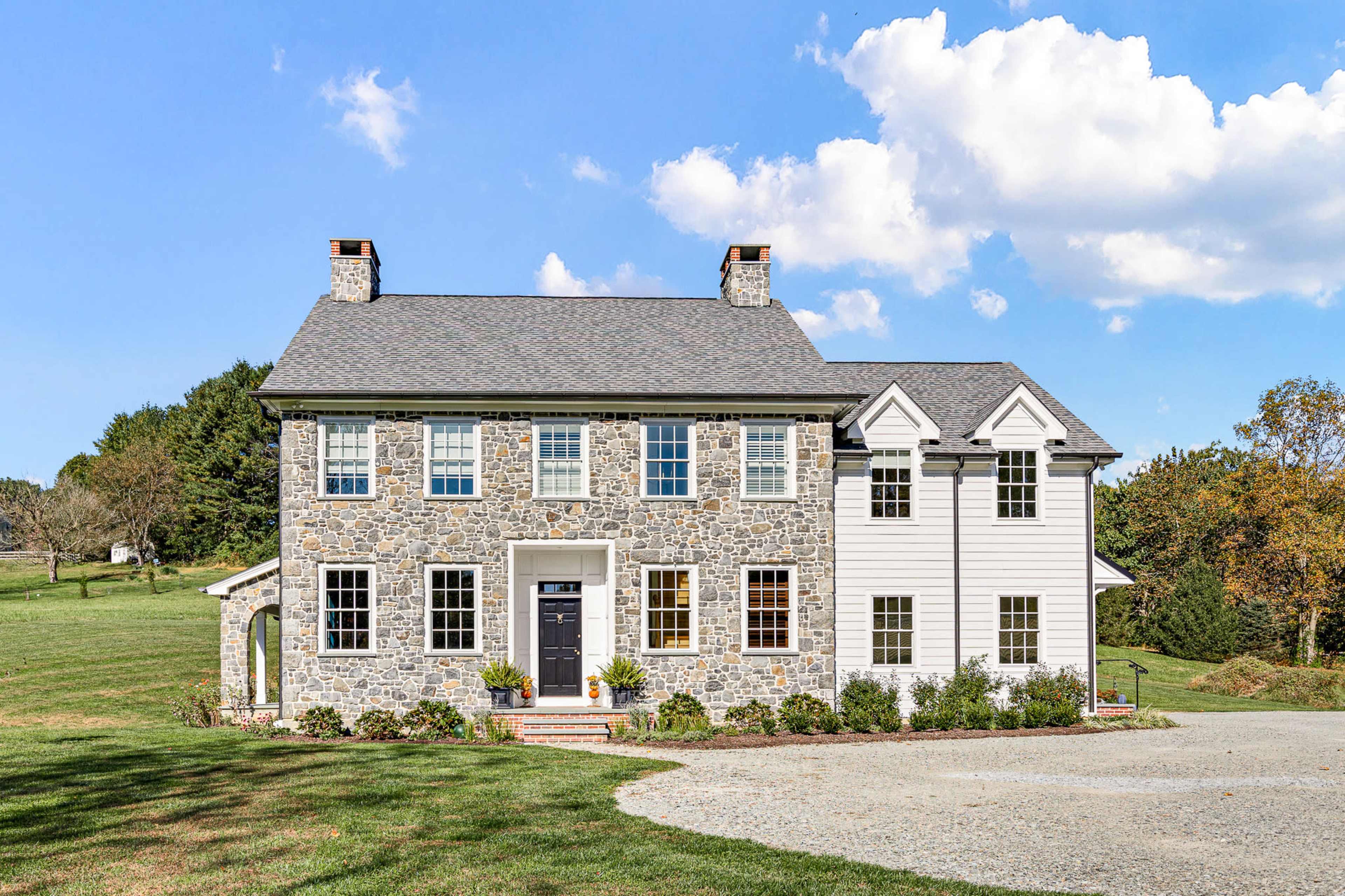 A two-story house features a stone exterior on the left side and white siding on the right, surrounded by a grassy yard and trees.