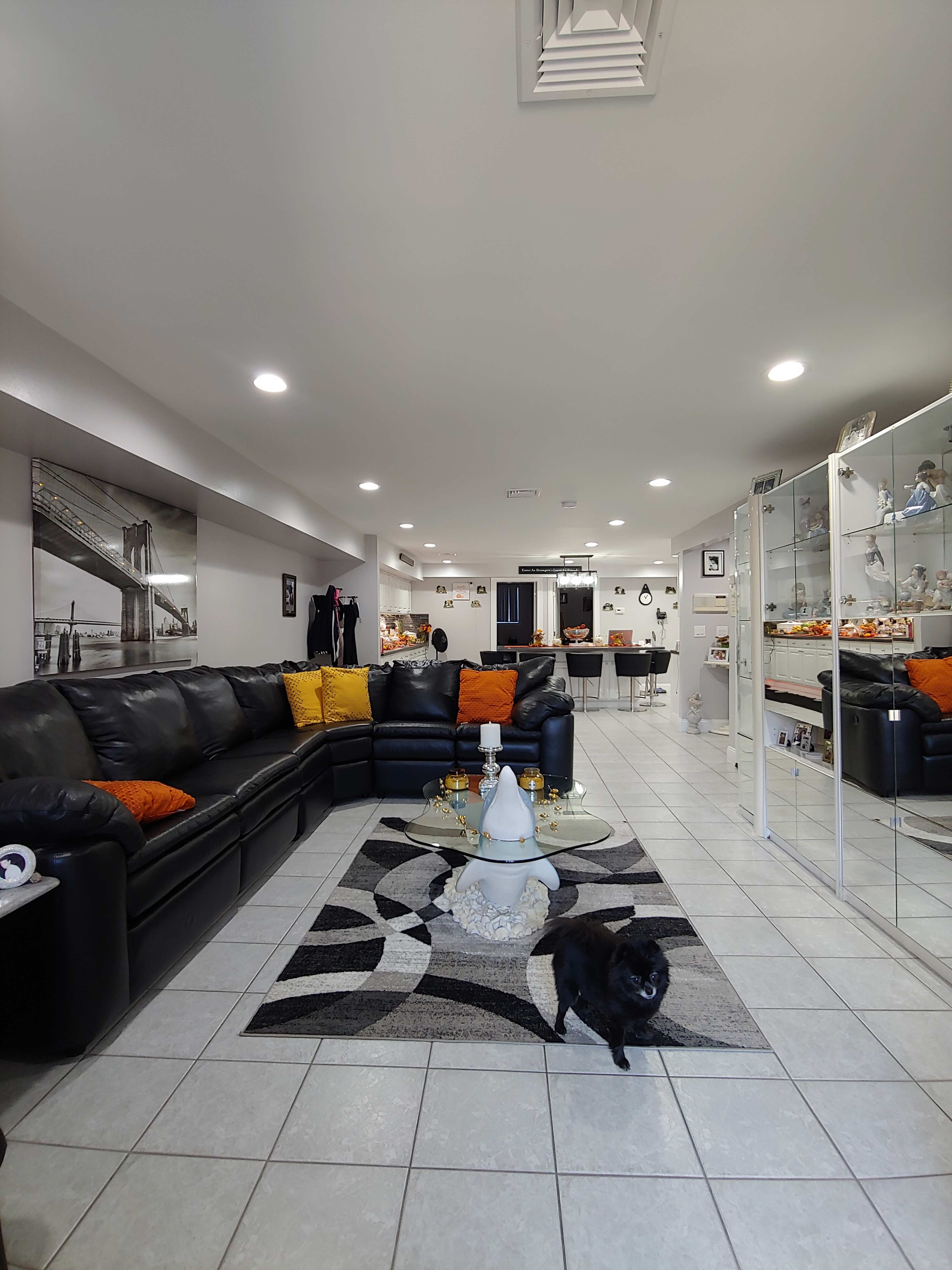 A modern basement living area with a black sectional couch, decorative pillows, a glass coffee table, and a small black dog on a tiled floor.