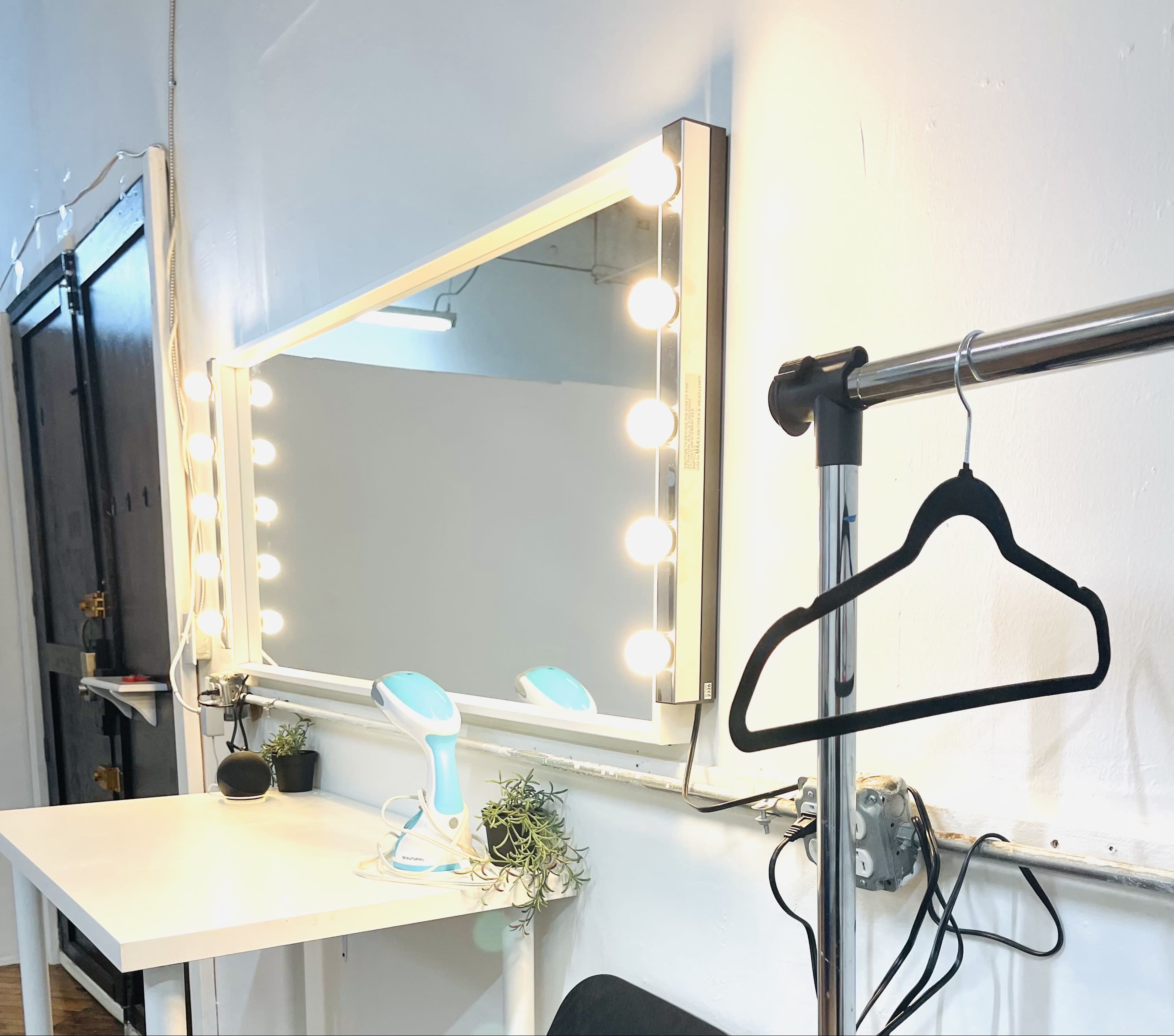 The image shows a well-lit vanity mirror with light bulbs, a clothes hanger on a pole, and a small ironing device on a table.