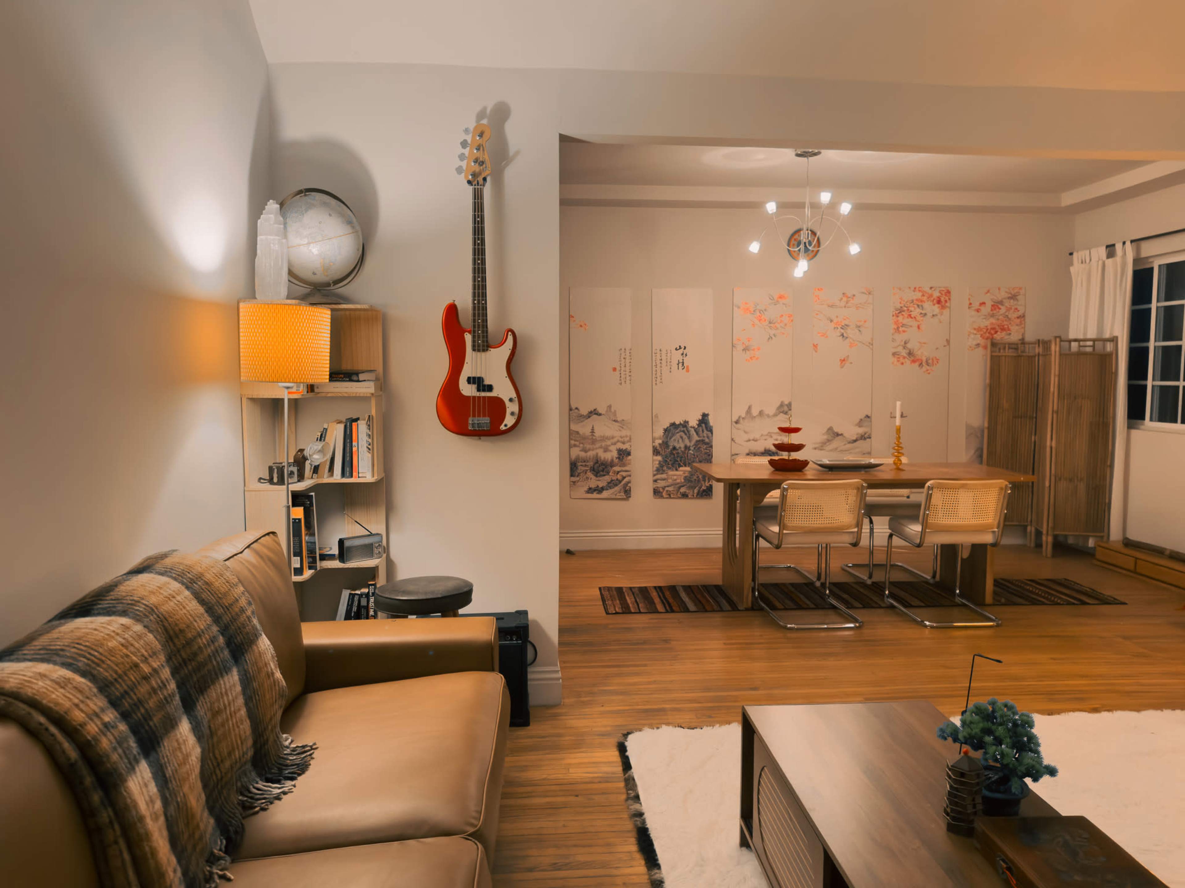 The image shows a cozy living room with a brown sofa and a red guitar hanging on the wall, leading to a dining area featuring a wooden table surrounded by chairs and decorative screens.
