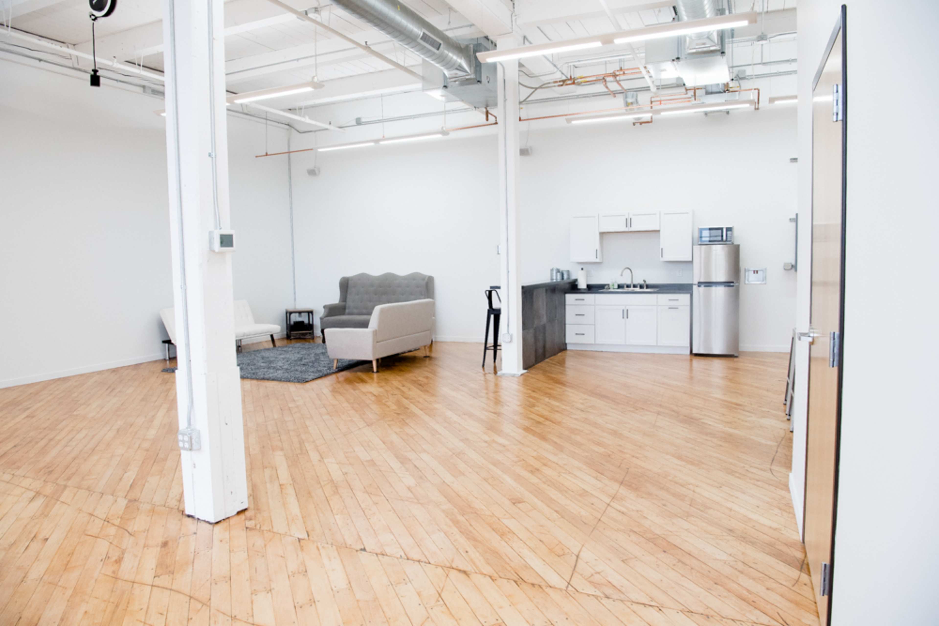 A spacious, minimalist interior featuring a wooden floor, a kitchen area with modern appliances, and a sofa set up alongside a rug.