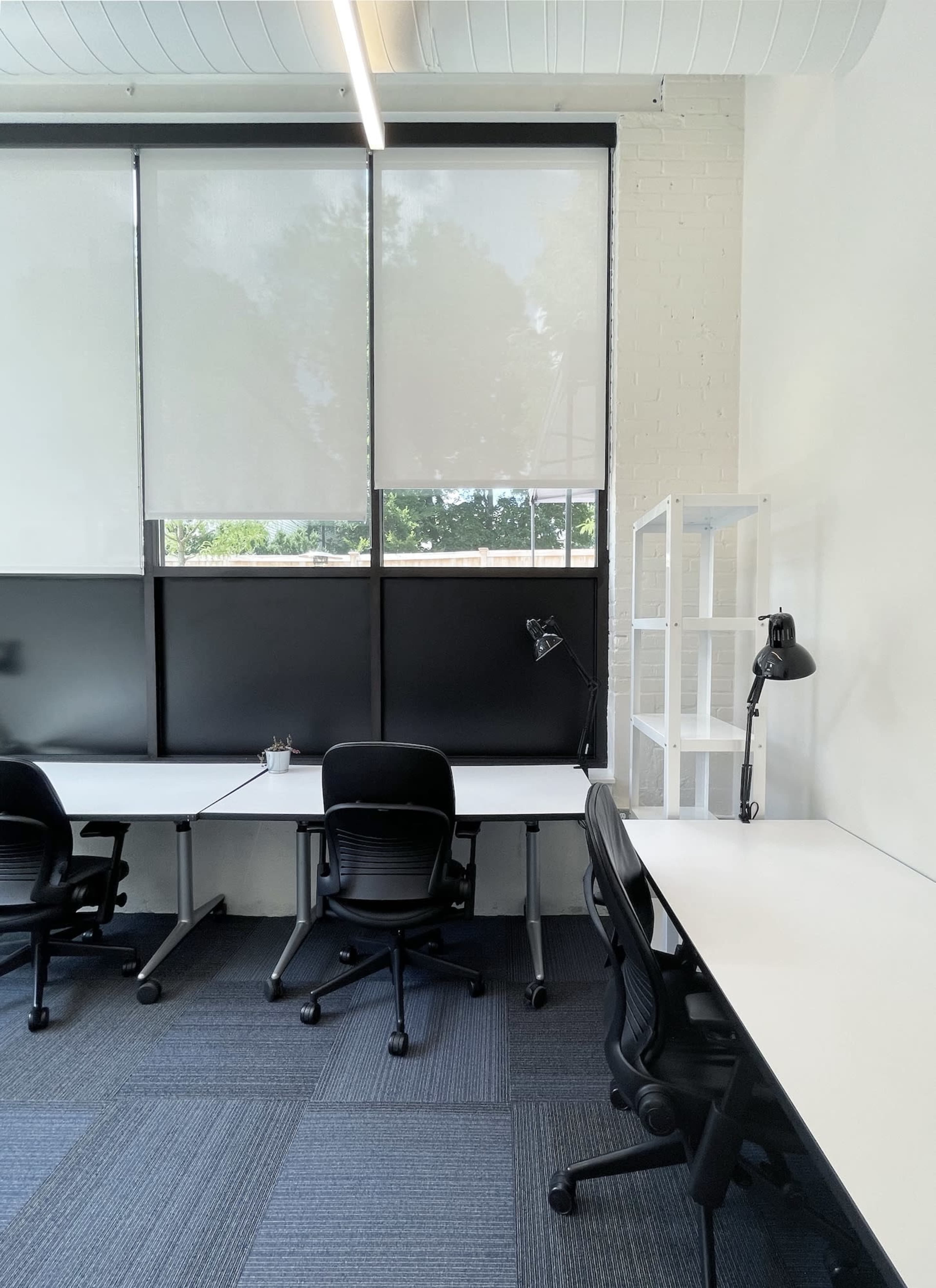 An office workspace with two empty desks and ergonomic chairs, large windows covered by light-filtering shades, and a tall shelving unit in the corner.