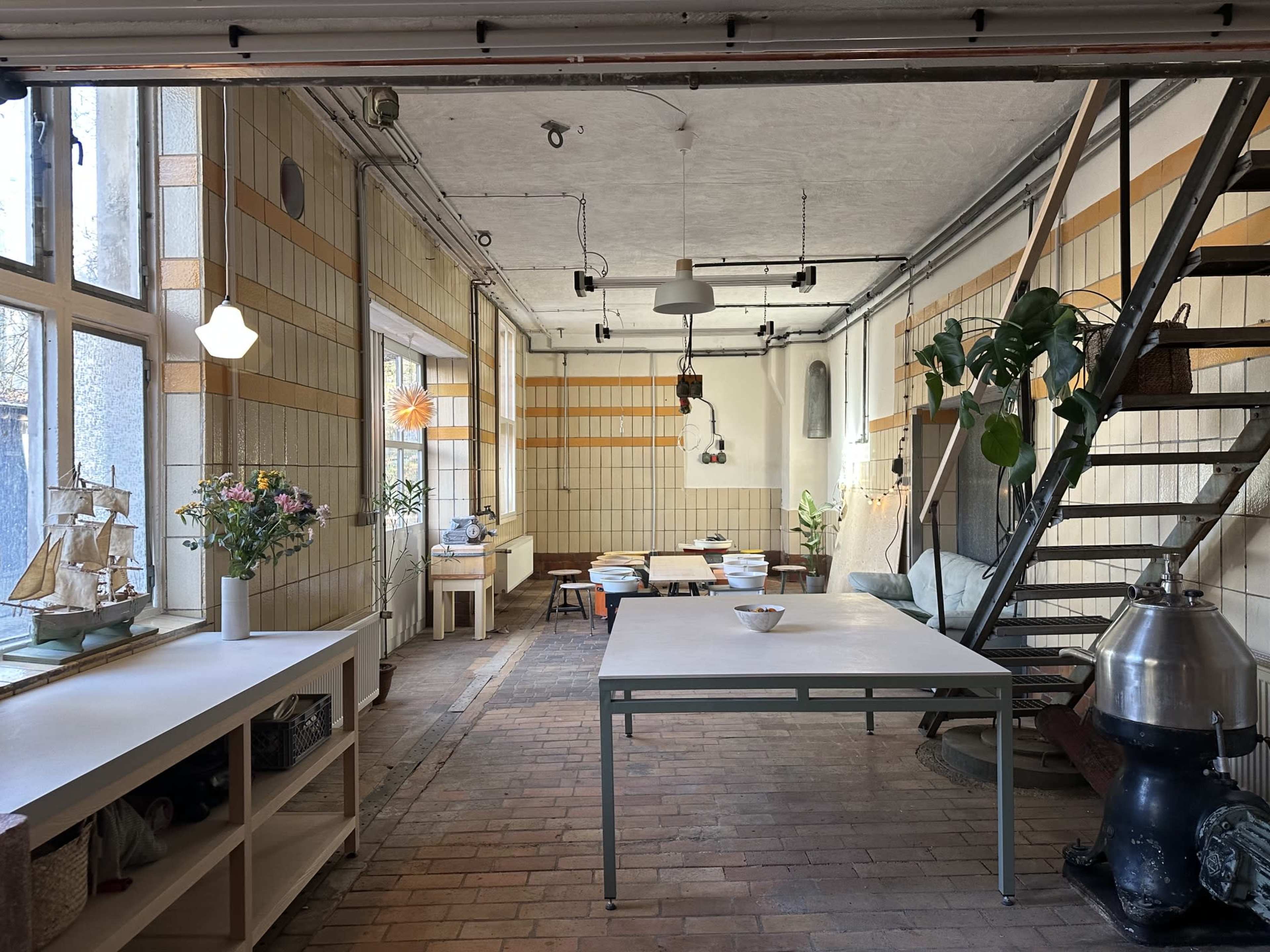 The image shows a spacious, well-lit room with a mix of dining and seating areas, featuring wooden floors, plants, and a staircase along one wall.