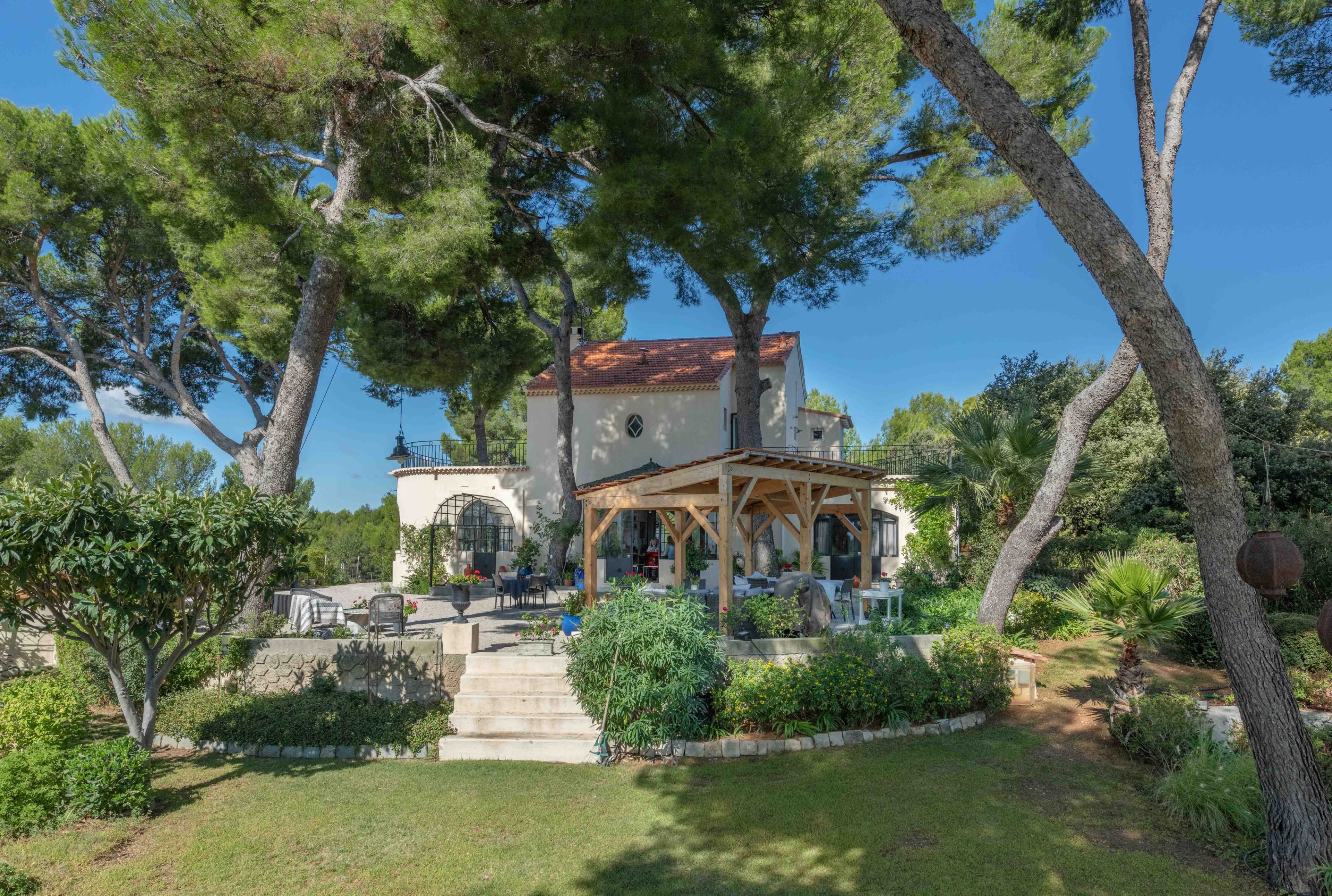 A house with a red-tiled roof is surrounded by tall trees and greenery, with a patio area in front and stone steps leading to the garden.