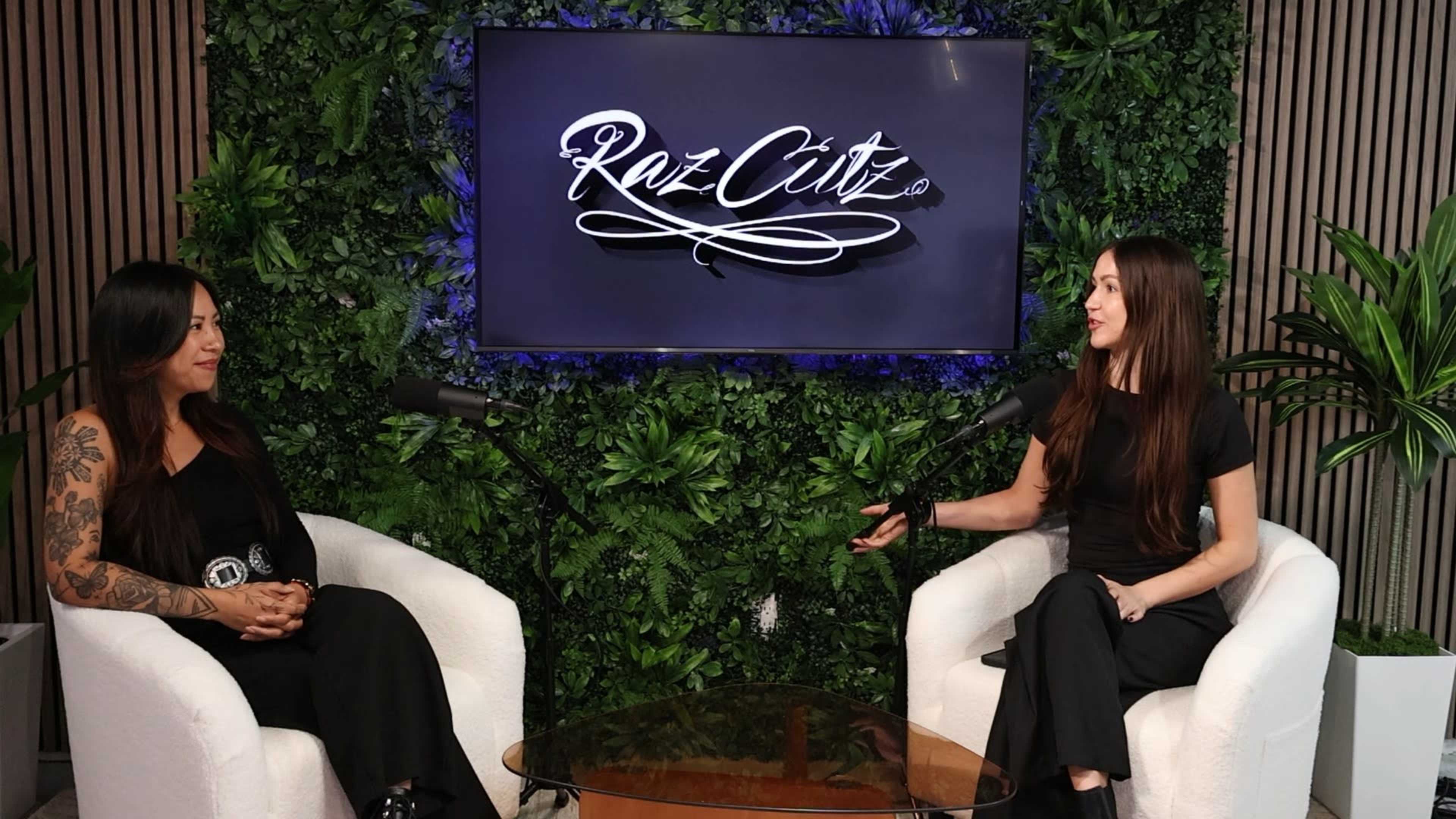Two women are seated on white chairs in front of a large screen displaying the logo "Raz Cutz," surrounded by a green plant backdrop.