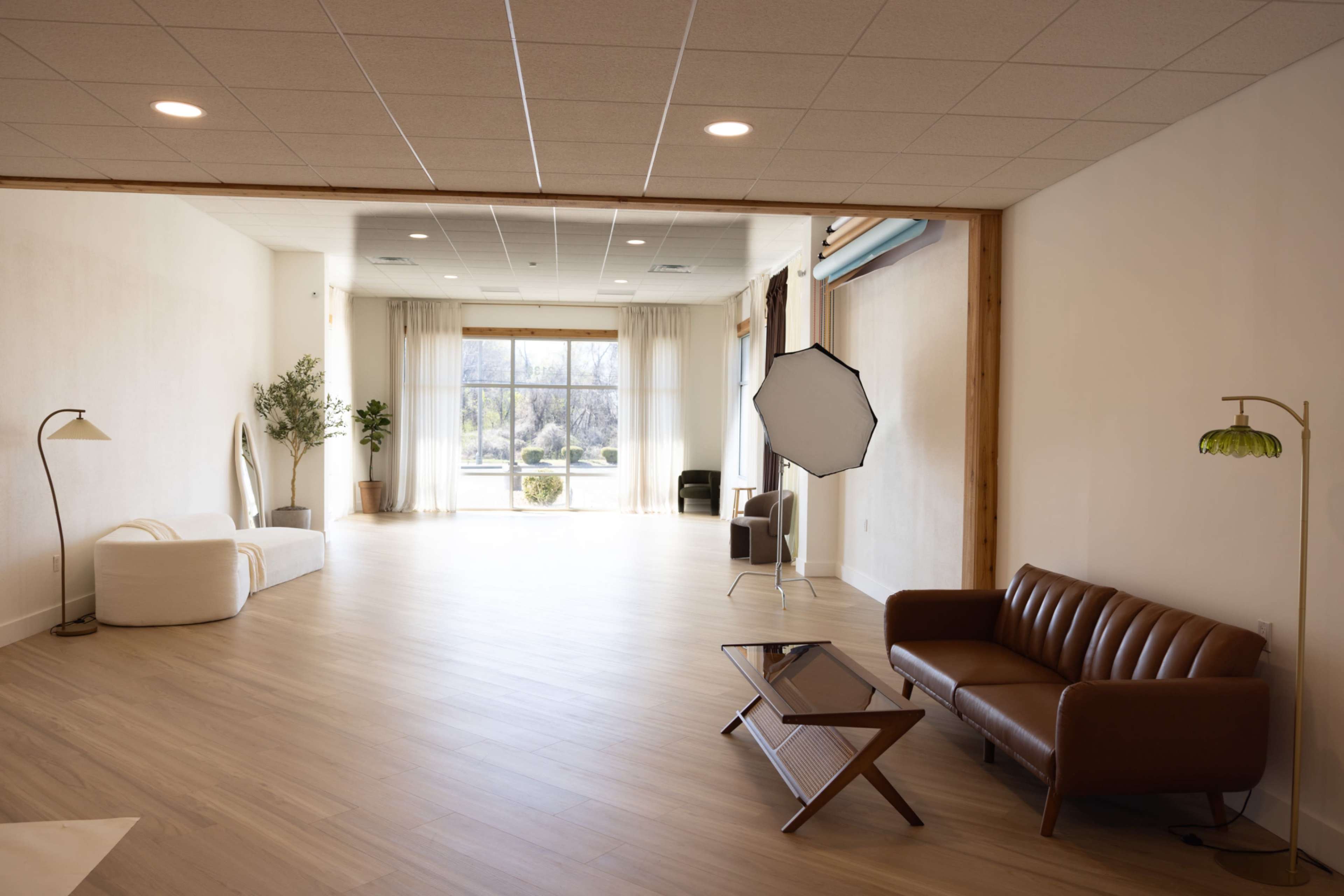 The image shows a spacious, well-lit room with wooden flooring, featuring a brown sofa, a coffee table, and large windows that let in natural light.
