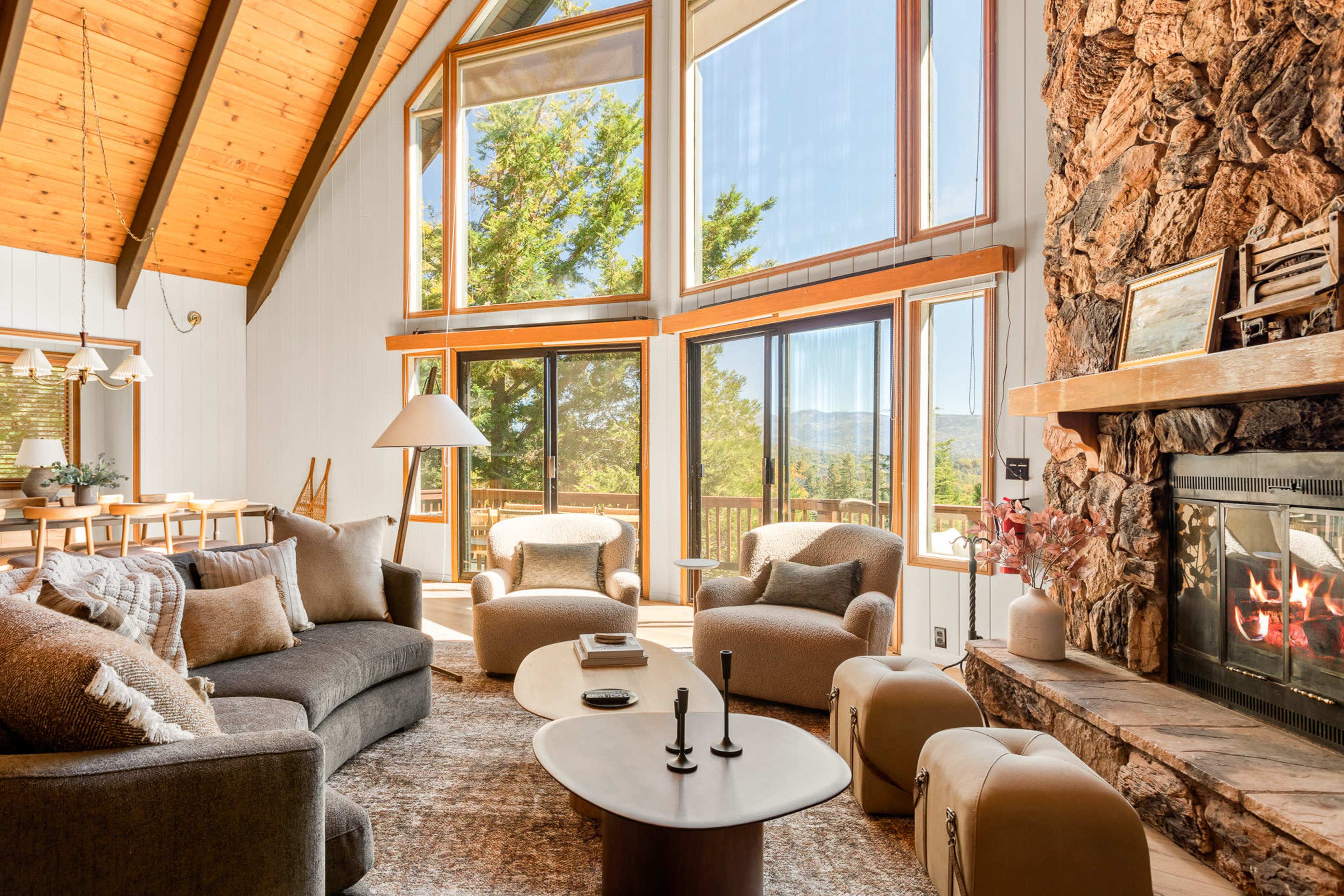 The image shows a spacious living room with large windows, a stone fireplace, and modern furniture arranged around a central table.