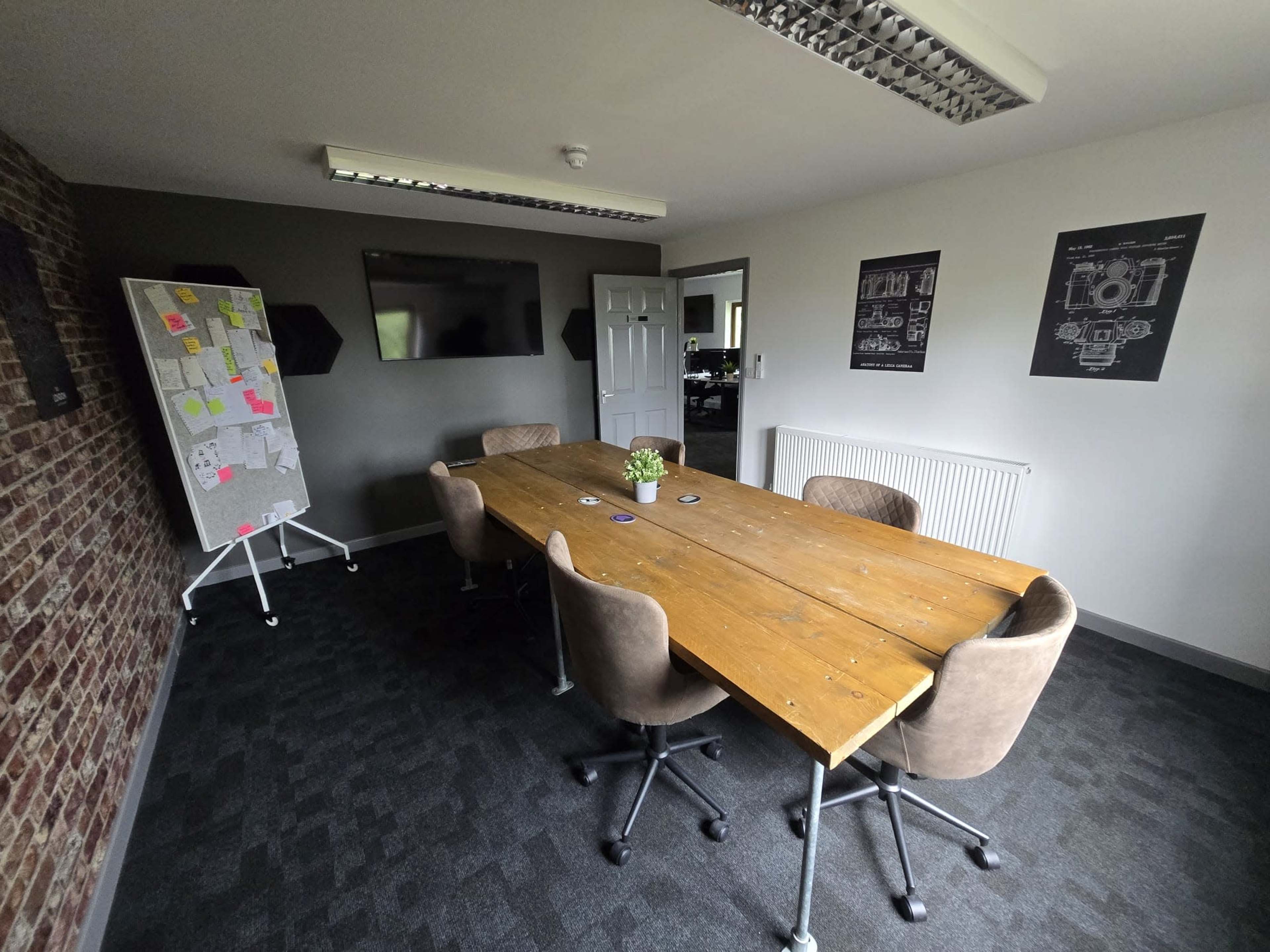 The image shows a modern conference room with a large wooden table surrounded by four chairs, a whiteboard with sticky notes, and a wall with posters.