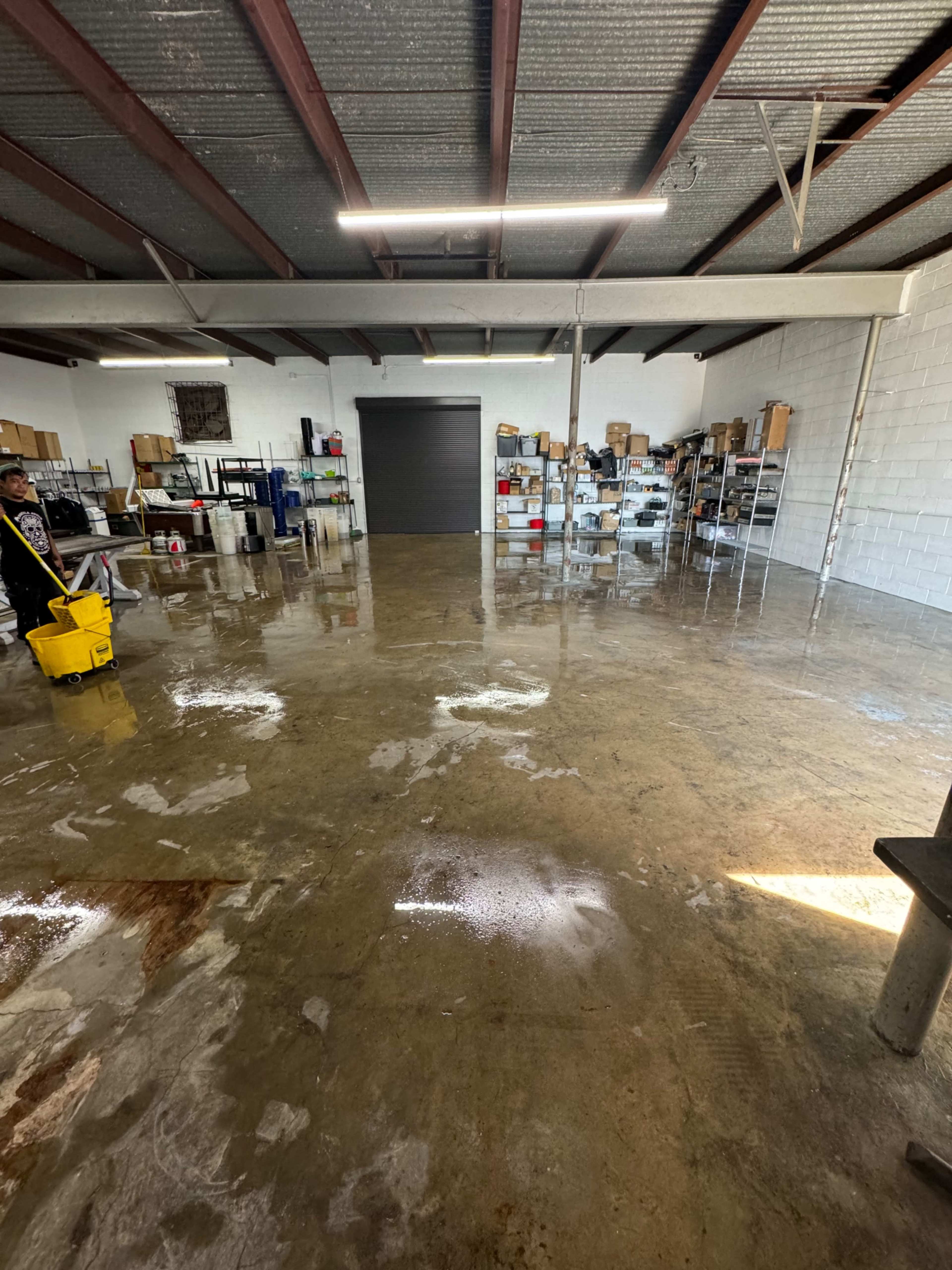 The image shows a clean, spacious garage or workshop with a polished concrete floor, shelves lined with boxes along the walls, and a person using a mop near a yellow bucket.