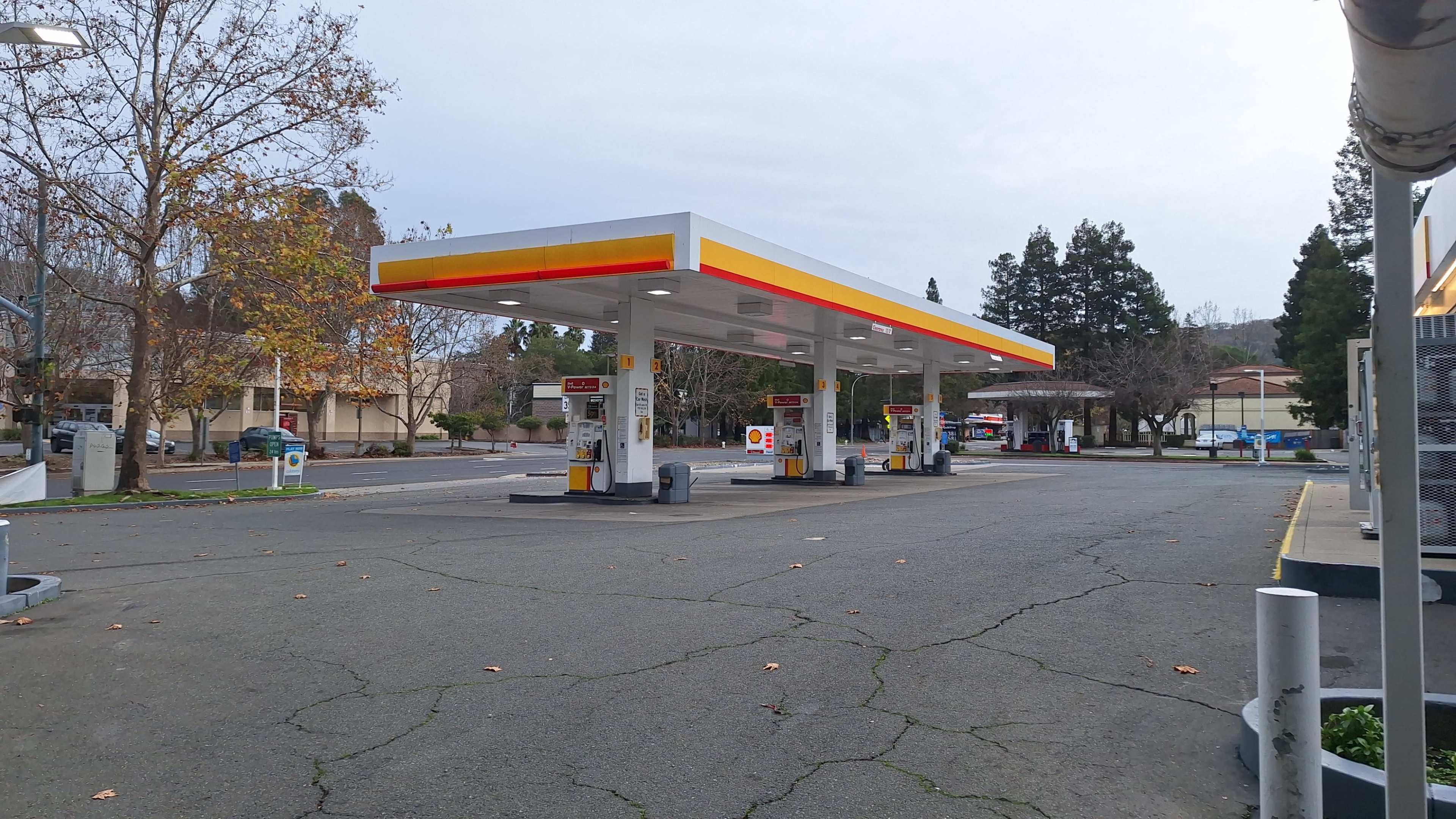 A Shell gas station with several fuel pumps is situated in a vacant parking lot surrounded by sparse trees and a few nearby buildings.