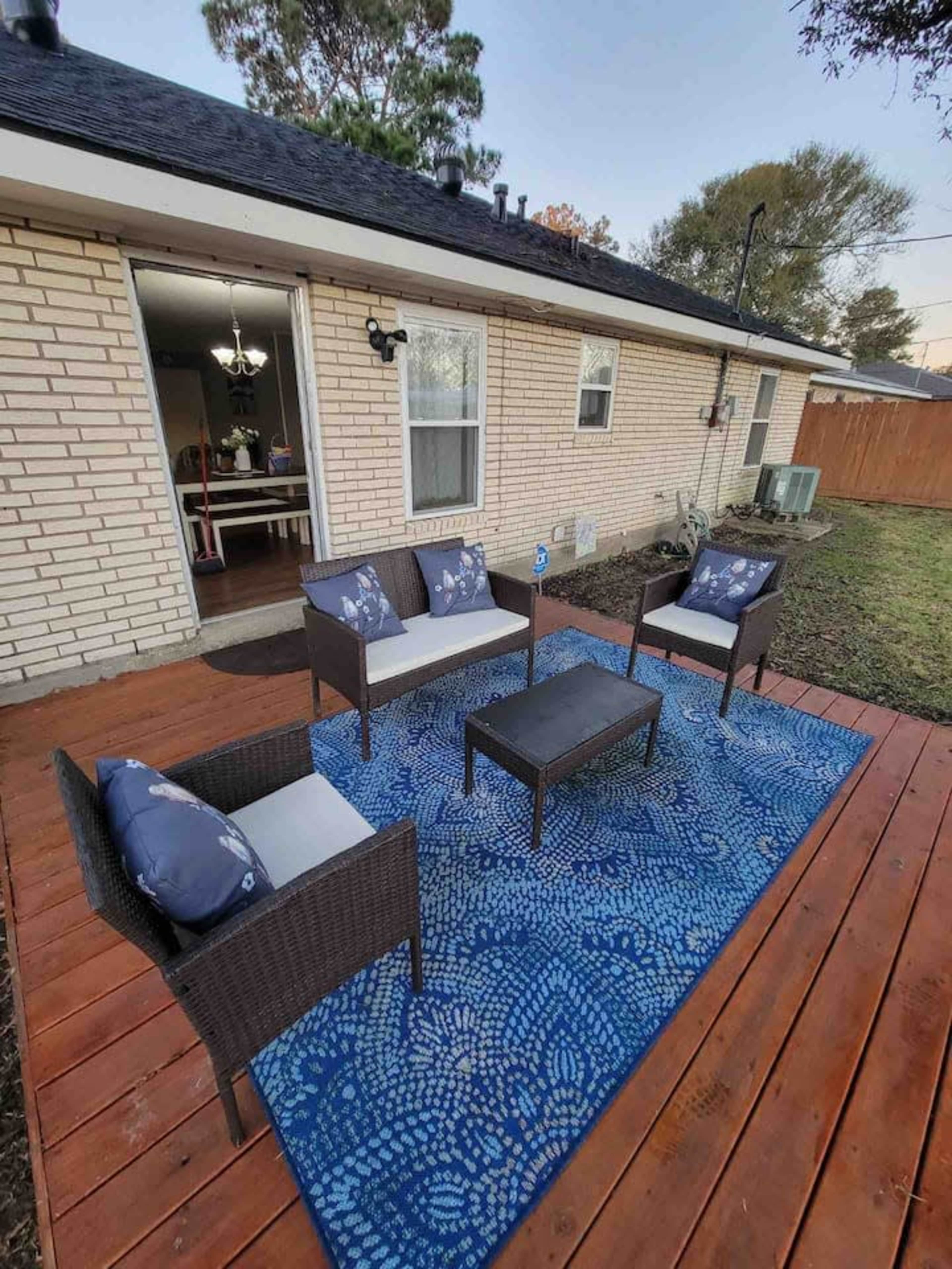 A patio area features four wicker chairs arranged around a small table on a blue patterned rug, with a house and grassy yard in the background.