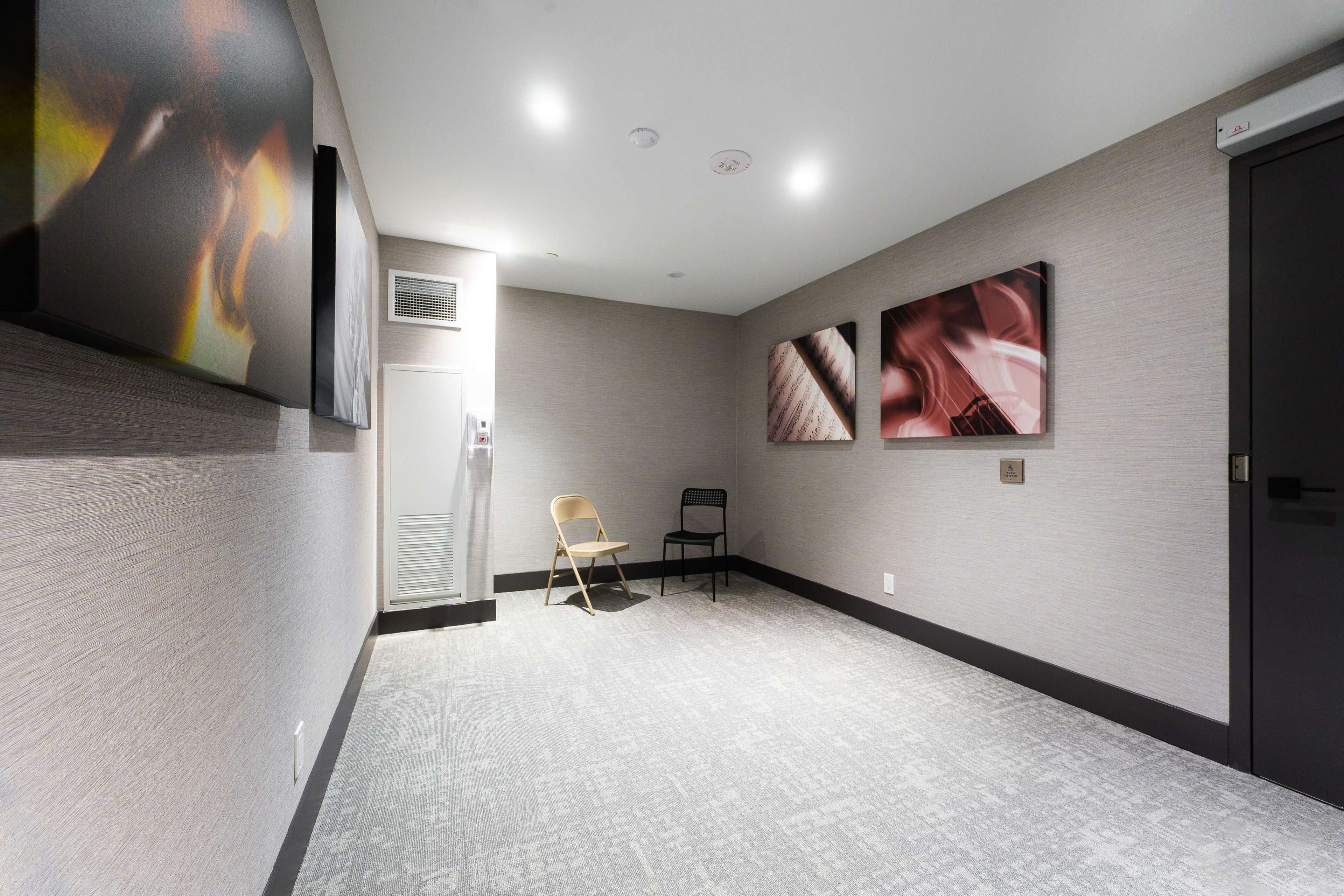 The image shows a minimalistic room with light gray walls, two abstract art pieces on the walls, and a folding chair positioned near the center.