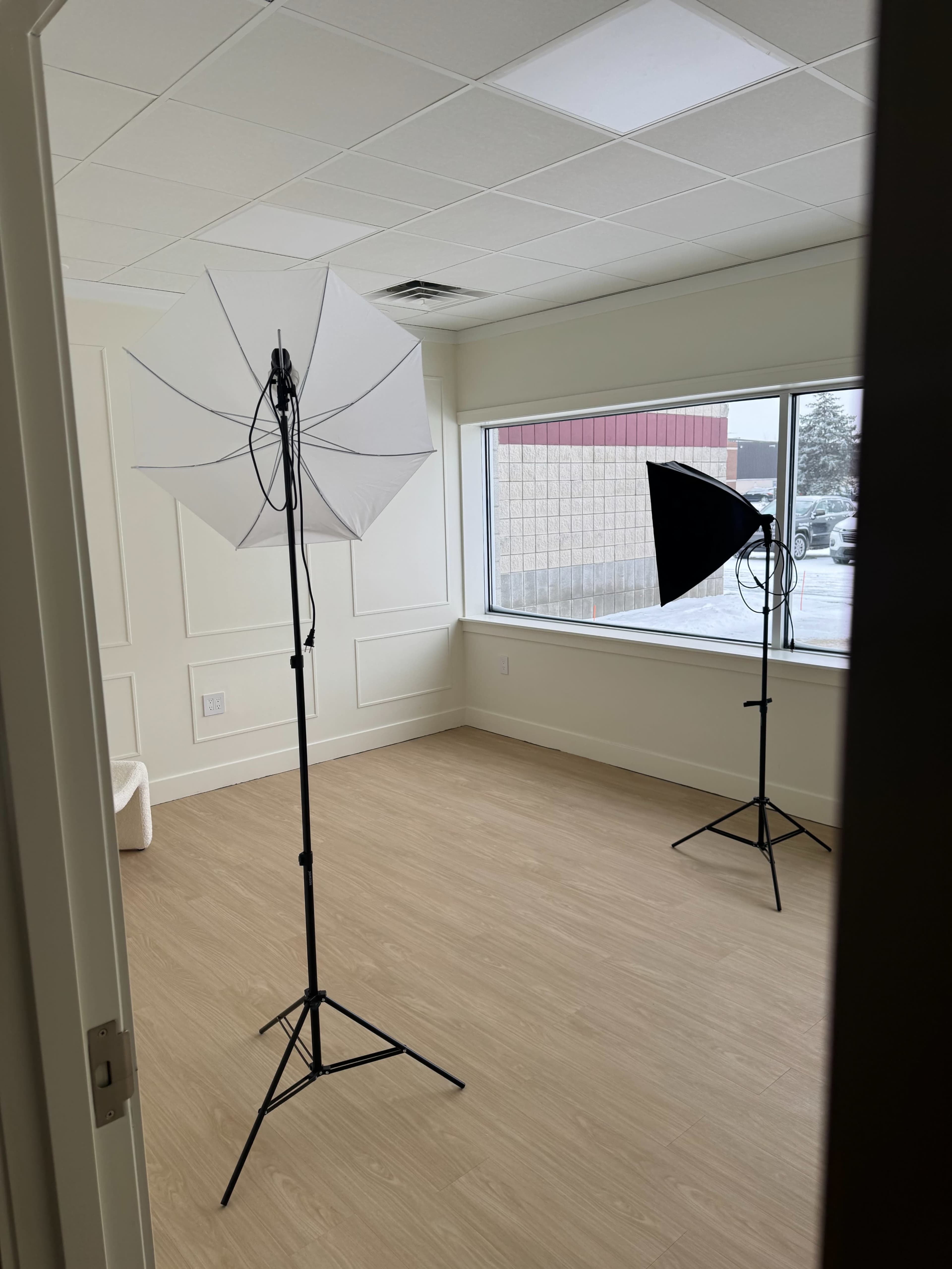 The image shows a minimalist room with two lighting umbrellas set up for photography, one white and one black, near a large window.